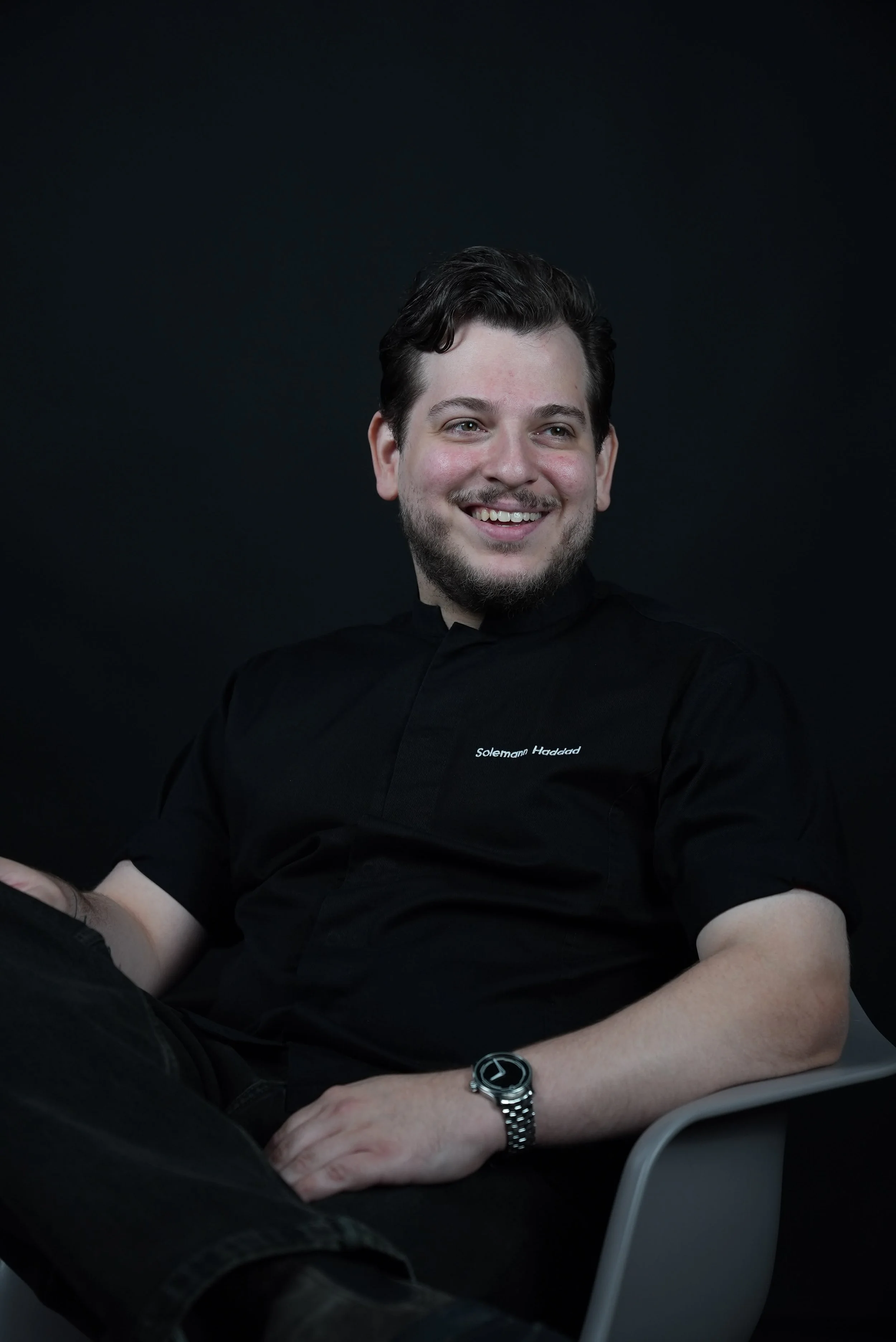 Young man with dark hair and beard smiling, wearing a black shirt with white text, seated at a desk with a black background, wearing a watch on his left wrist.