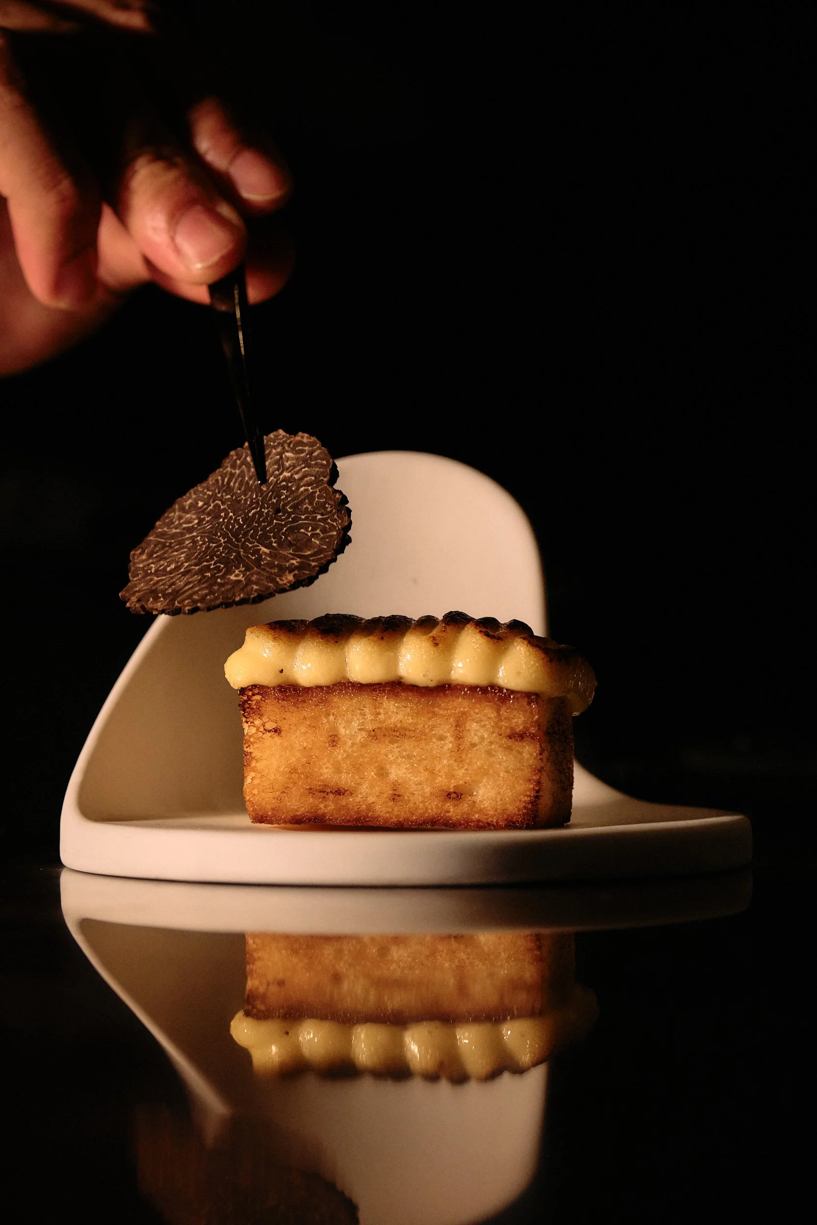 Close-up of a dessert with a rectangular cake topped with a layer of cream and a toasted, browned crust, placed on a white plate. A small piece of black truffle or chocolate garnishes the cake. The background is black, with the plate and dessert refl