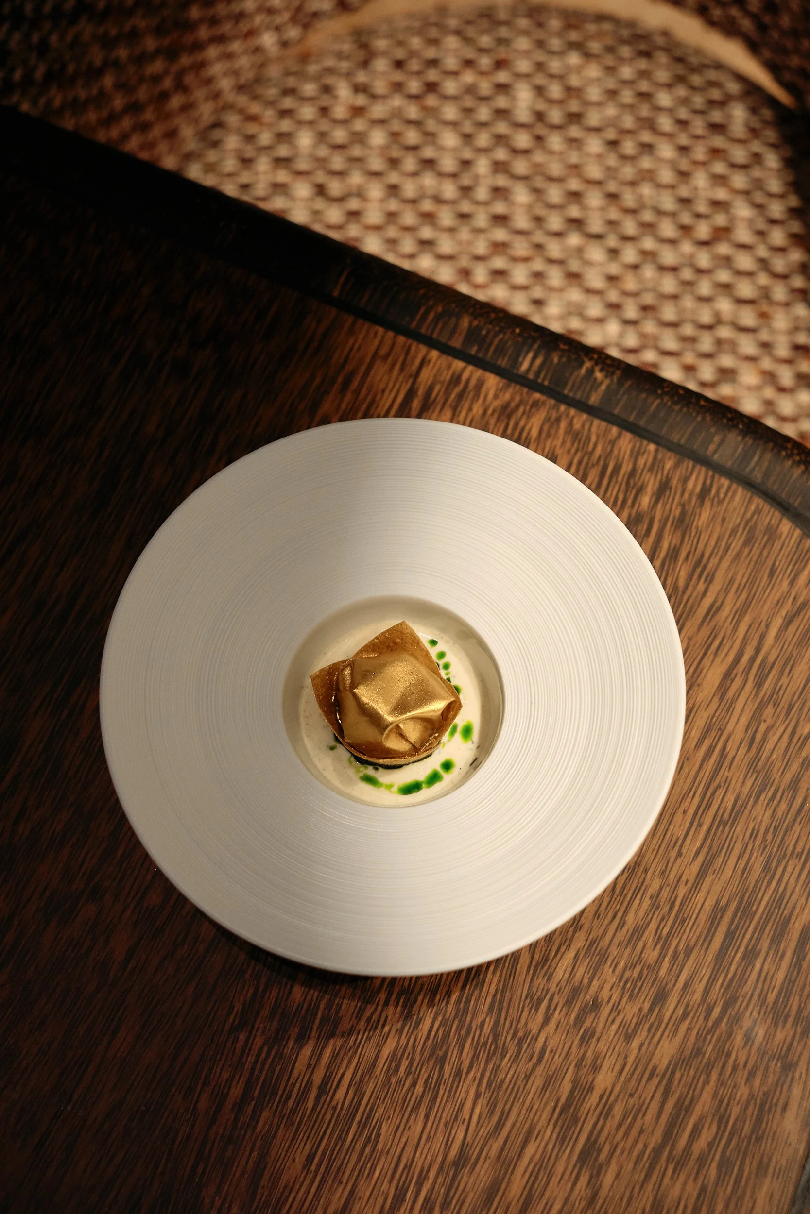 A white plate with a square piece of dessert wrapped in gold-colored foil, garnished with green sauce dots around it, on a dark wooden table.