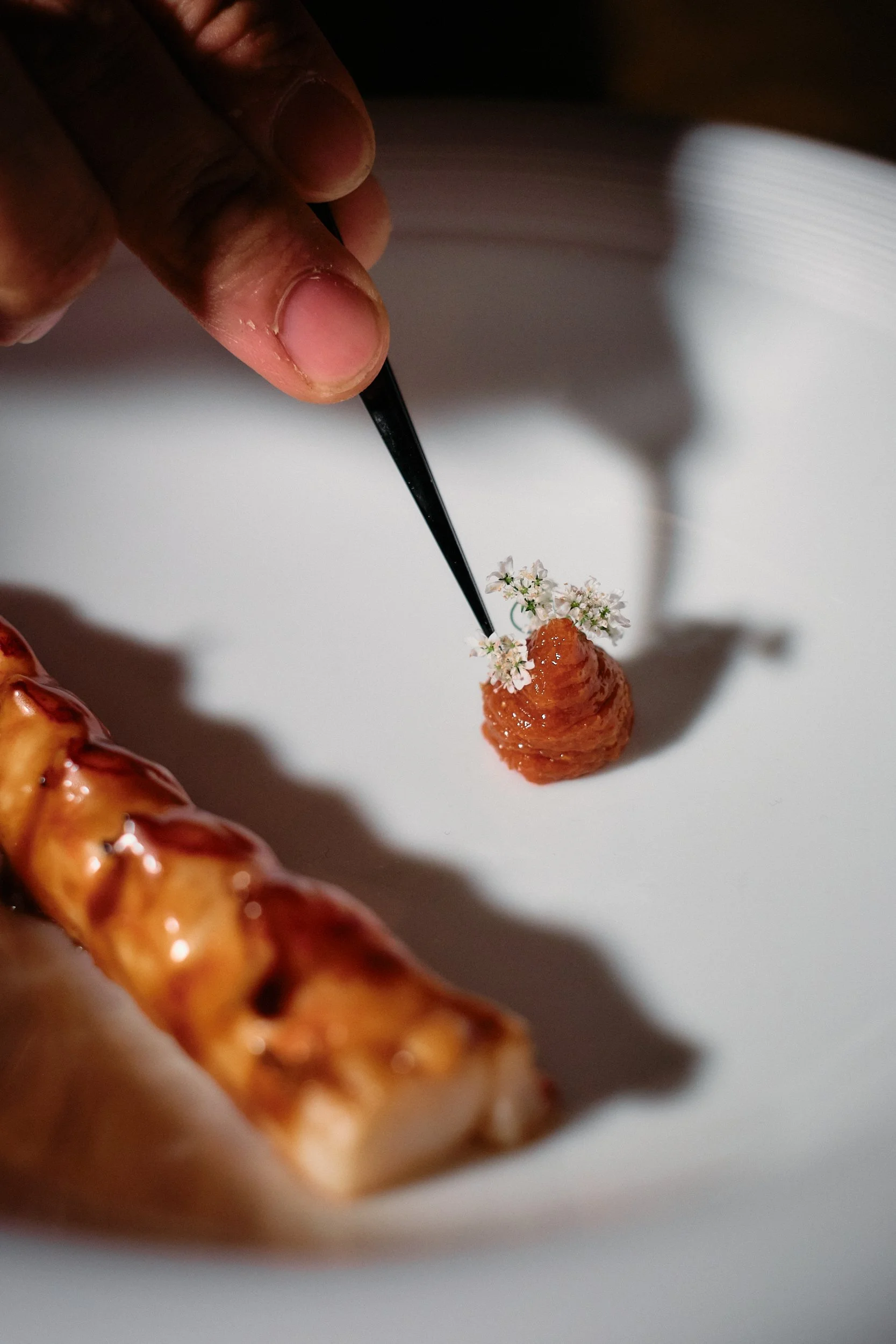 Close-up of a hand holding a thin black tool with a small white flower, orange fruit, or pastry, on top of a white surface, with part of a pizza seen in the lower left corner.