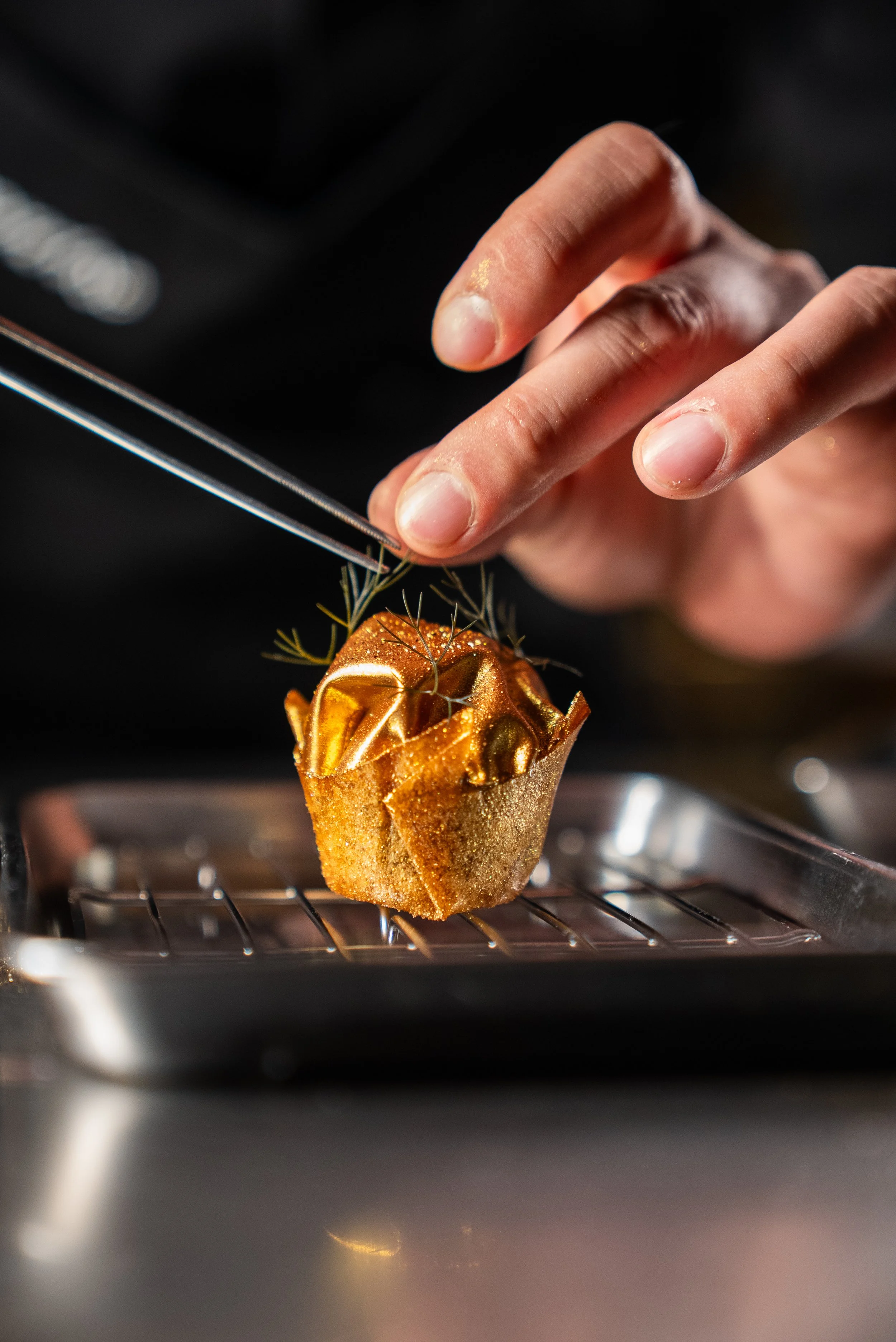 Mans hand placing a garnish on a gold nugget with a tweezer in his hands. 