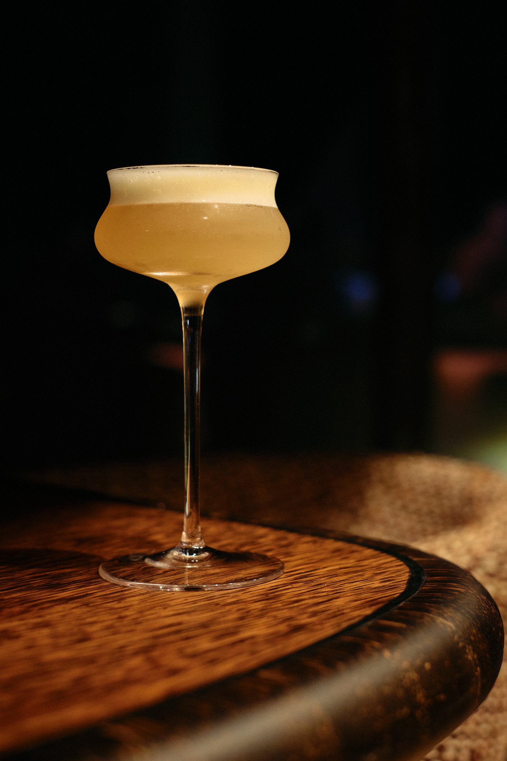 A cocktail in a stemmed glass with a frothy top, placed on a wooden surface with a dark background.