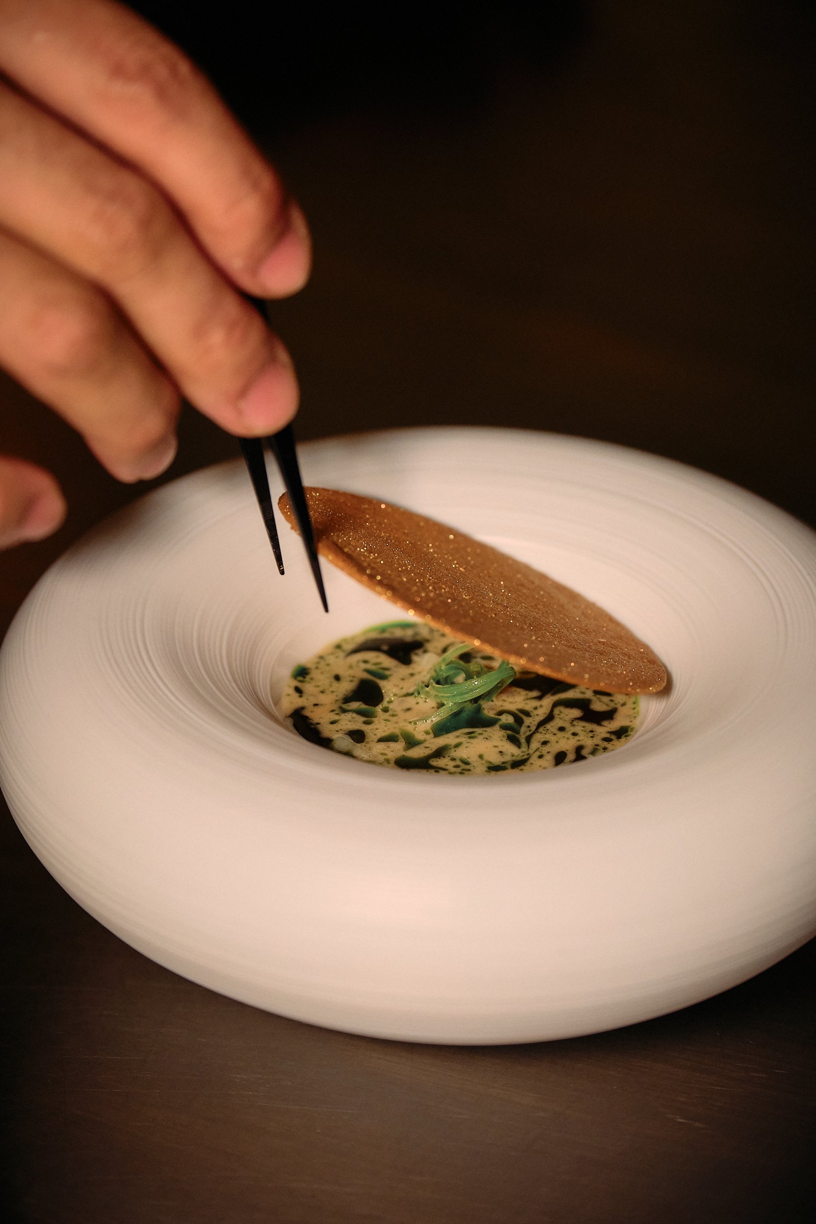 A hand holding chopsticks picks up a piece of food from a white bowl with a decorative rim, containing a creamy dish with green herbs and a drizzle of dark sauce, topped with a gold-colored cracker or crisp.