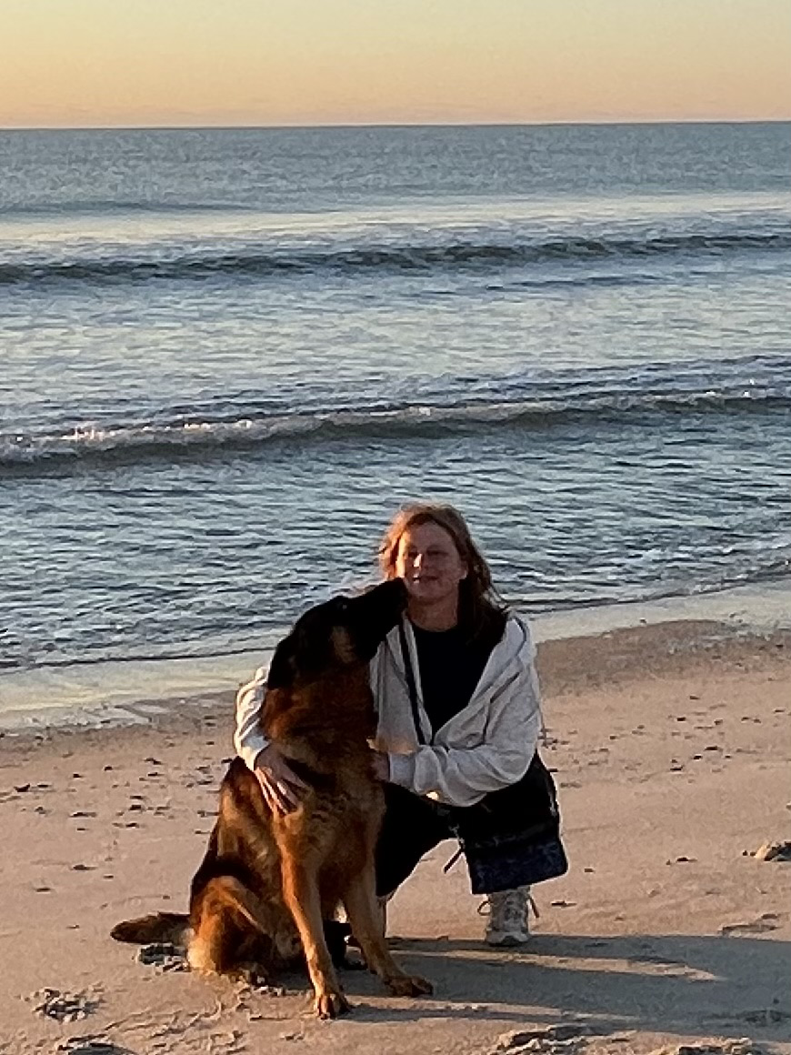 A woman and her large dog on a beach during sunset, with the ocean and sky in the background.