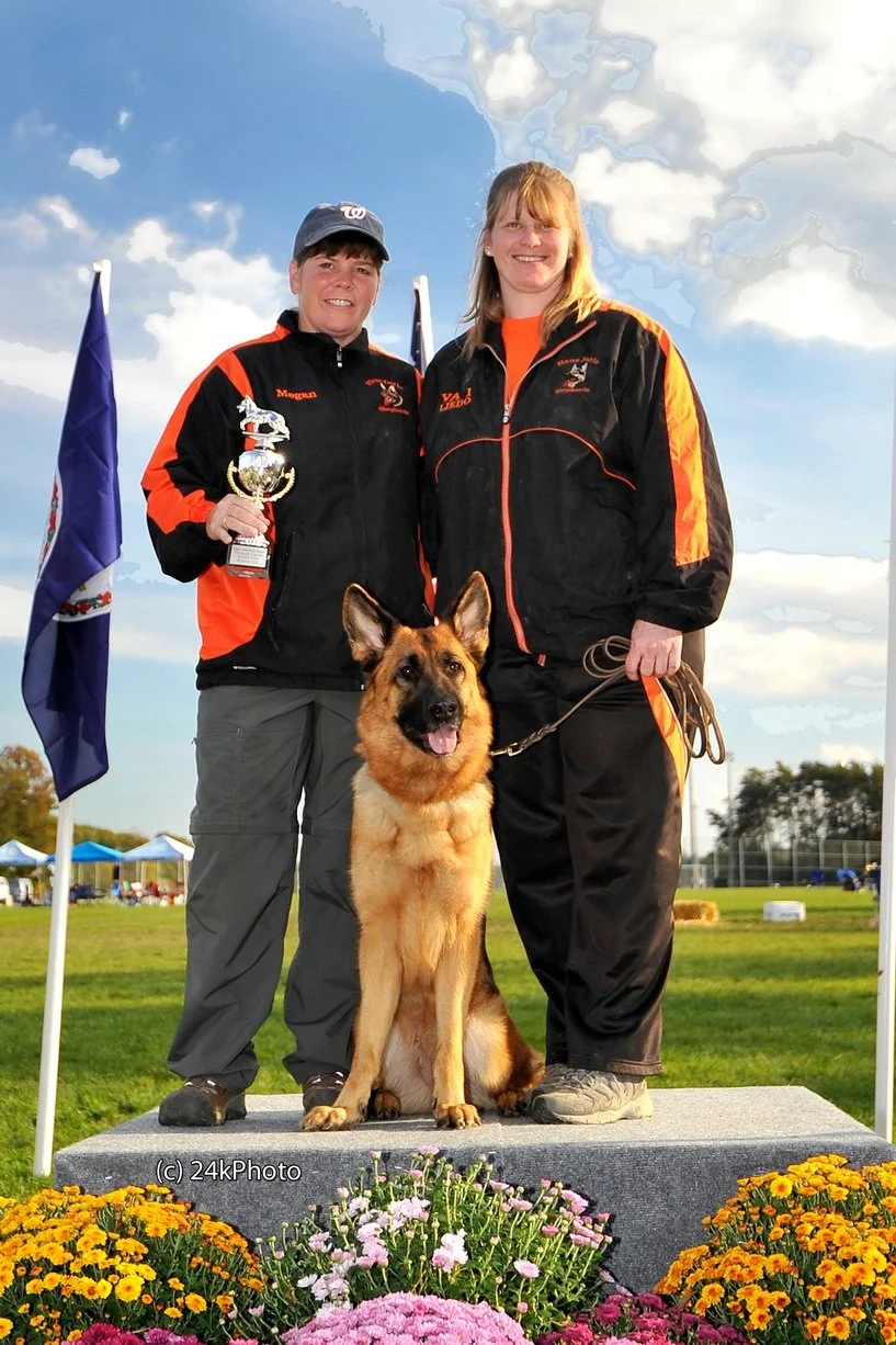 Two women in black and orange jackets standing outdoors on a podium with a German Shepherd dog and a woman holding a trophy amid a field with flowers, flags, and tents in the background.