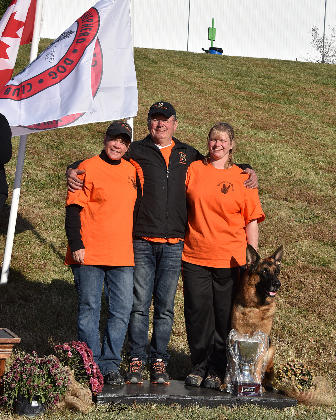 Three people standing outdoors on a grassy hill, smiling, with a German Shepherd dog sitting in front of them. They are dressed in orange shirts and black jackets or pants, and two are wearing caps. Flowers and a trophy are on the ground in front of them.