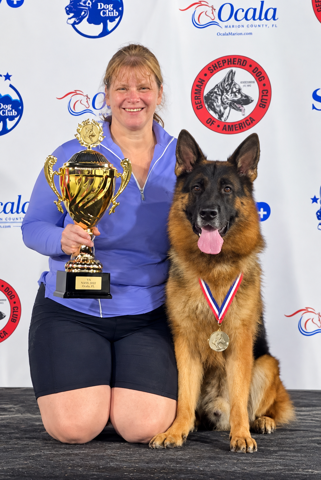 A woman kneeling on her knees holding a large gold trophy, next to a German Shepherd wearing a gold medal with a red, white, and blue ribbon. The background displays logos of dog clubs and organizations, indicating a dog competition award ceremony.