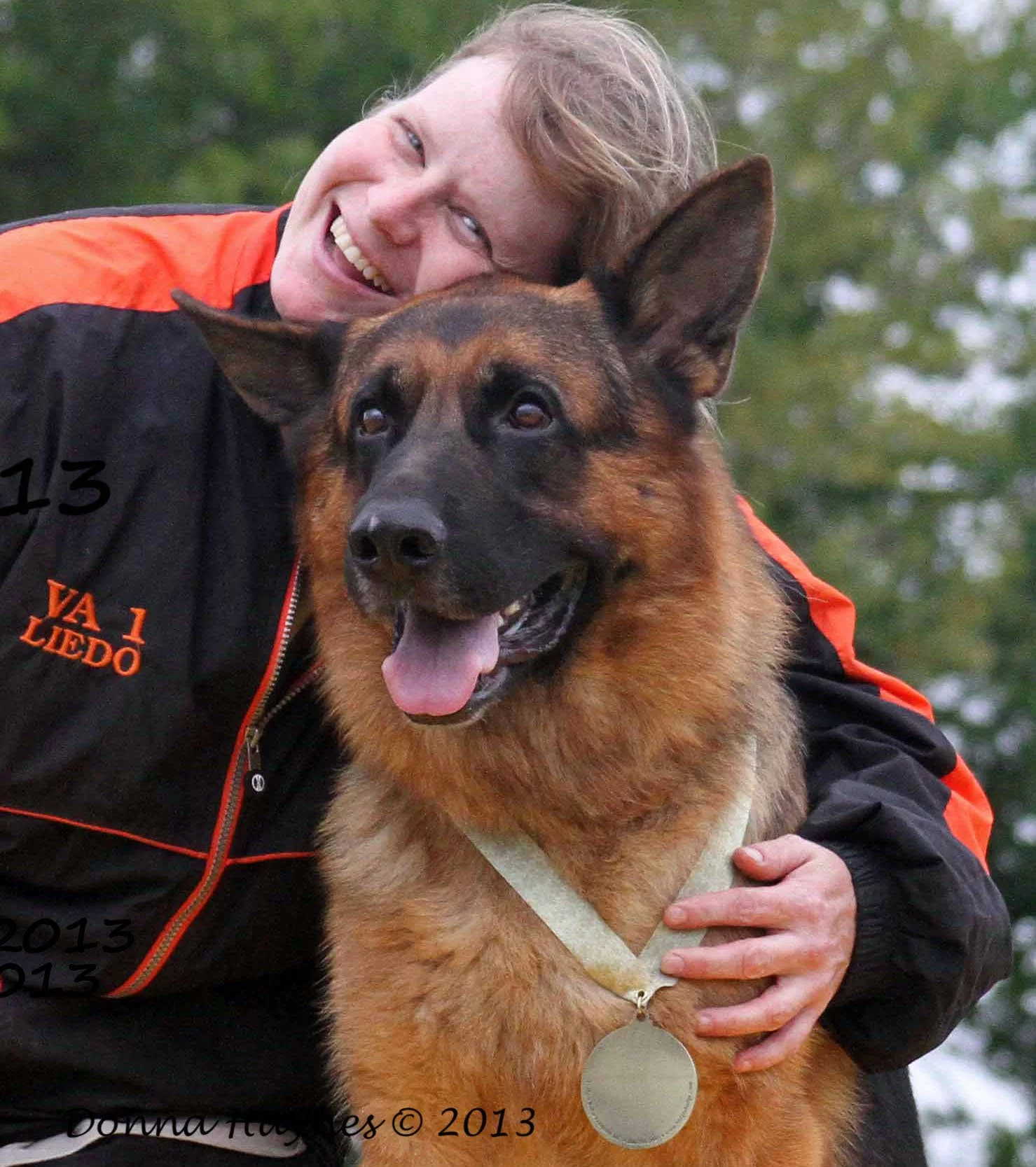 A woman with short, curly hair smiling and hugging a German Shepherd dog during daytime. The woman is wearing a black and orange jacket, and the dog wears a large medal around its neck. Trees are blurred in the background.