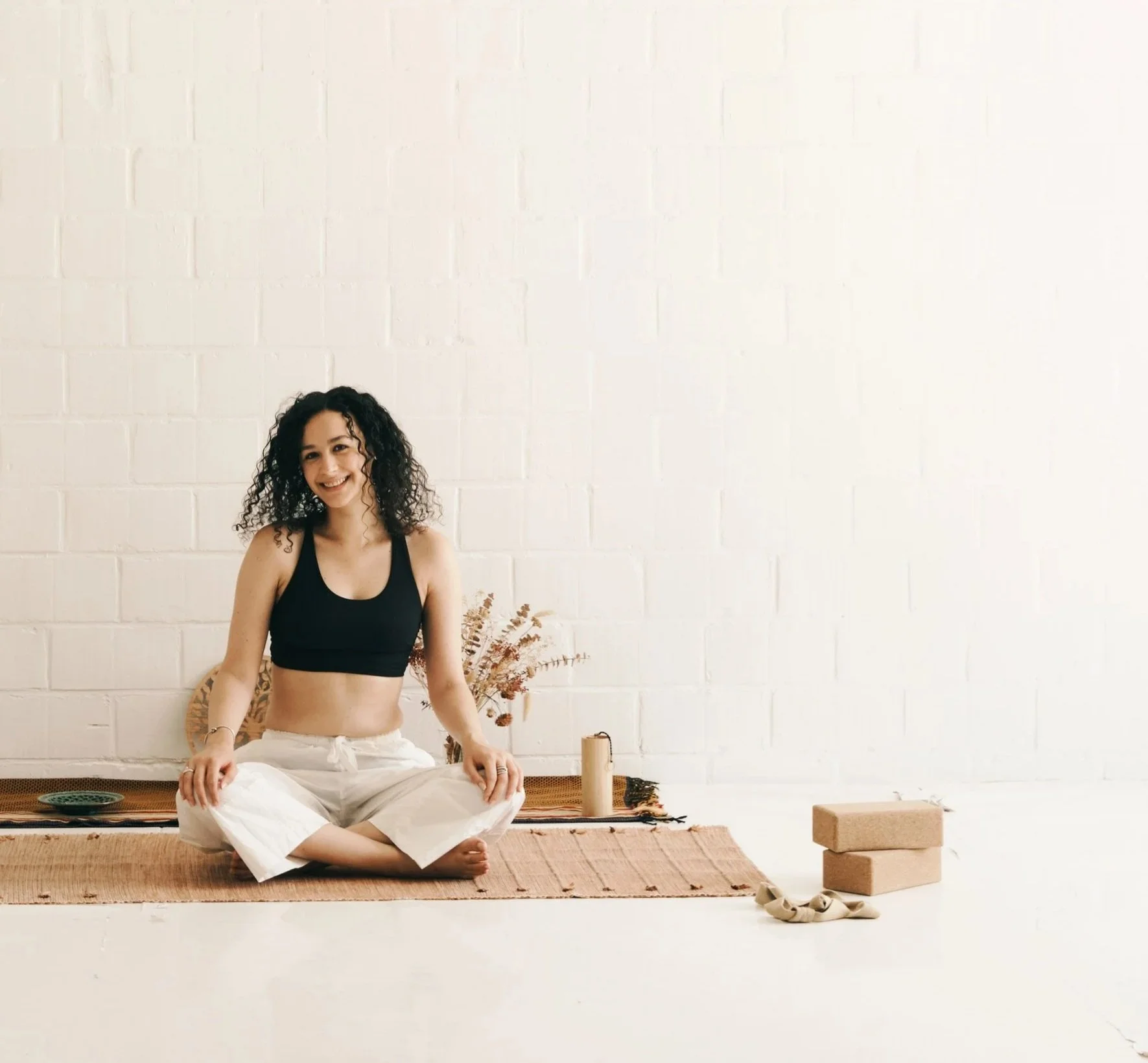 femme souriante assise en tailleur sur un tapis de yoga. blocs de yoga, instrument de musique et fleurs séchées pour décorer l'espace. Accueil chaleureux, douceur et sérénité.
