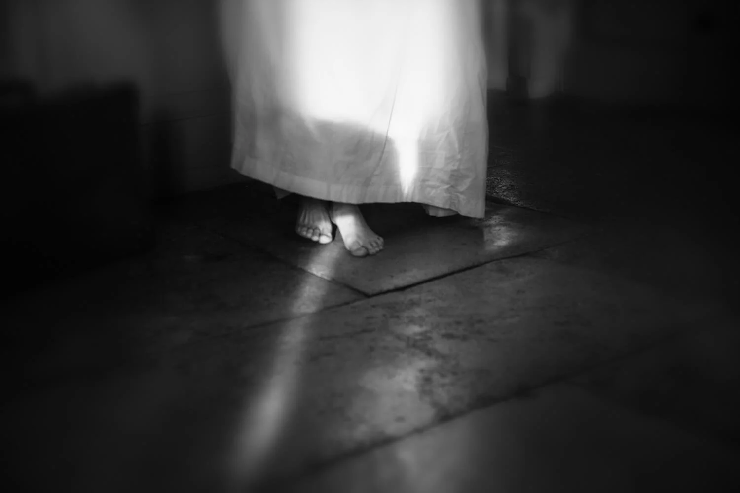 Detail of Bare Feet by Ania Ready, showing bare feet and the hem of a white dress illuminated across a dark floor.