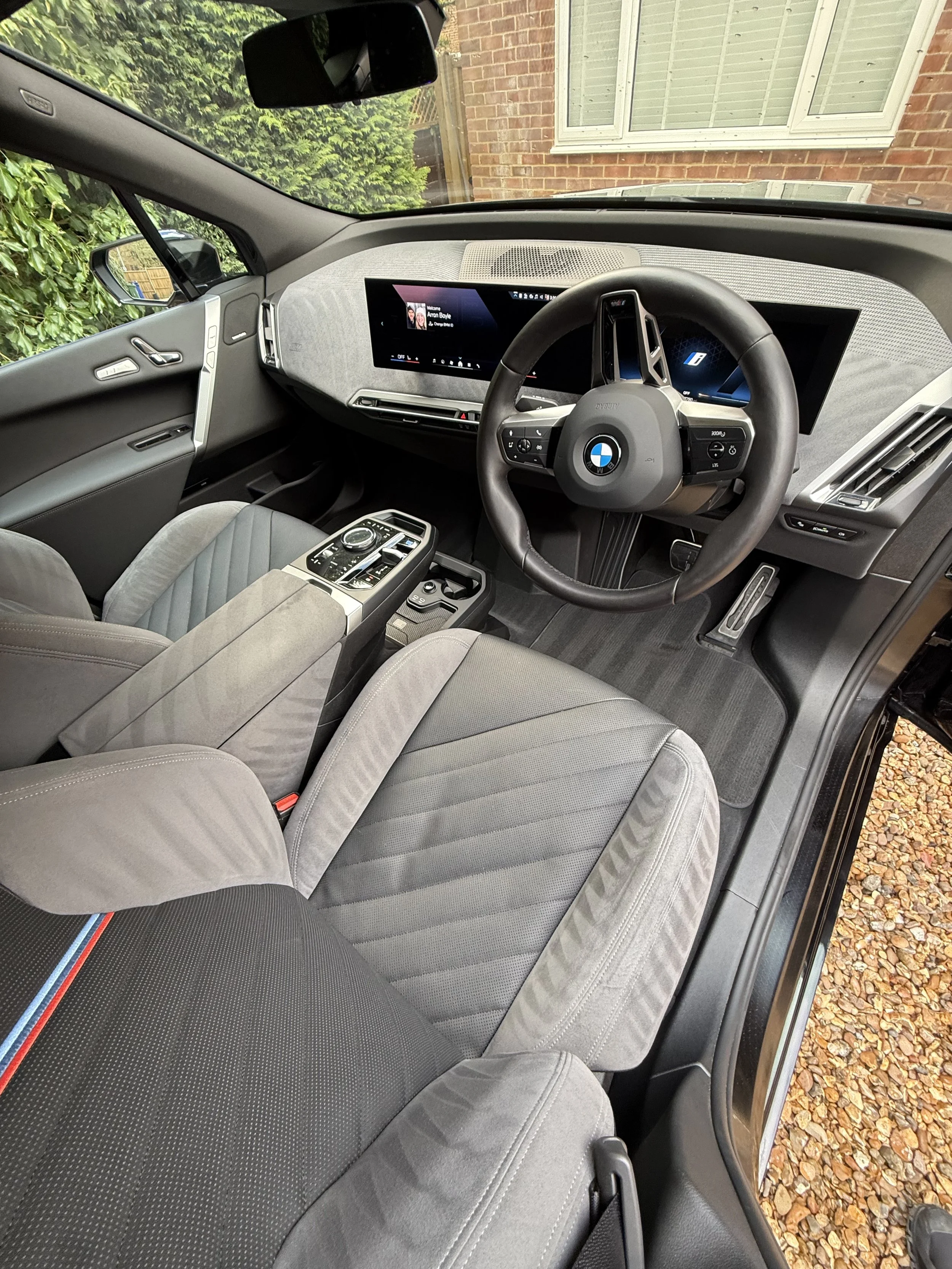The interior of a modern BMW vehicle showing the dashboard, steering wheel, and front seats, with a view of the driver's side door and outdoor gravel ground.