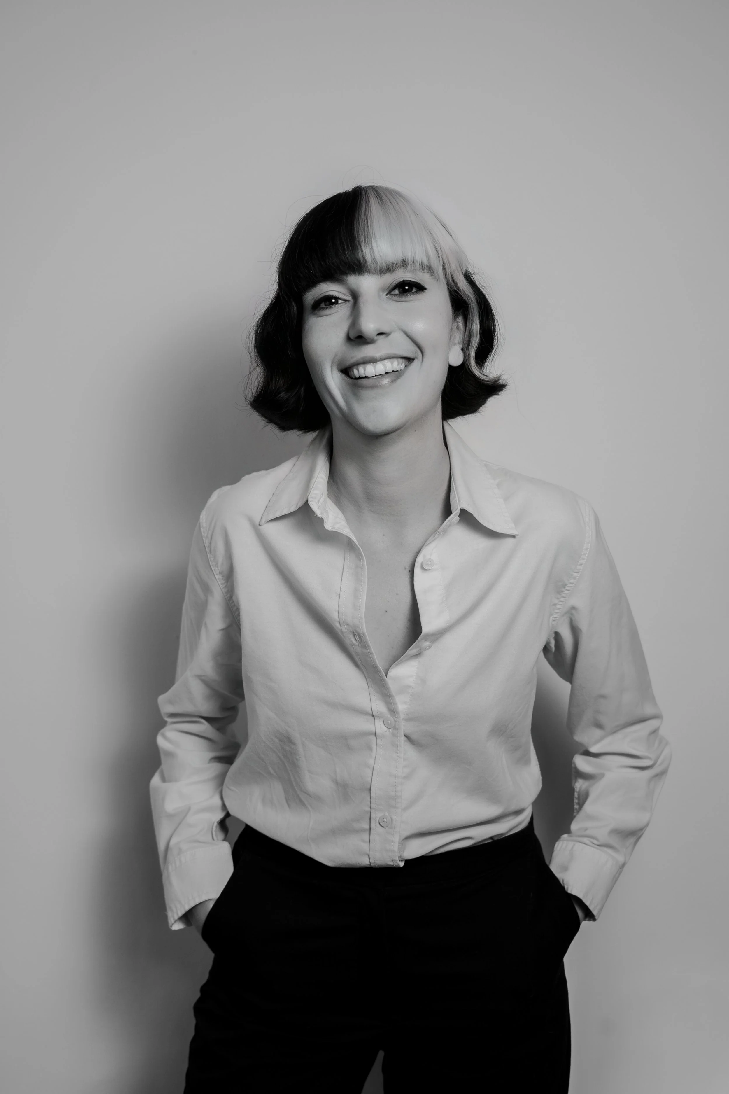 Black and white photo of a woman smiling, wearing a light-colored button-up shirt and dark pants, standing with hands on hips against a plain background.