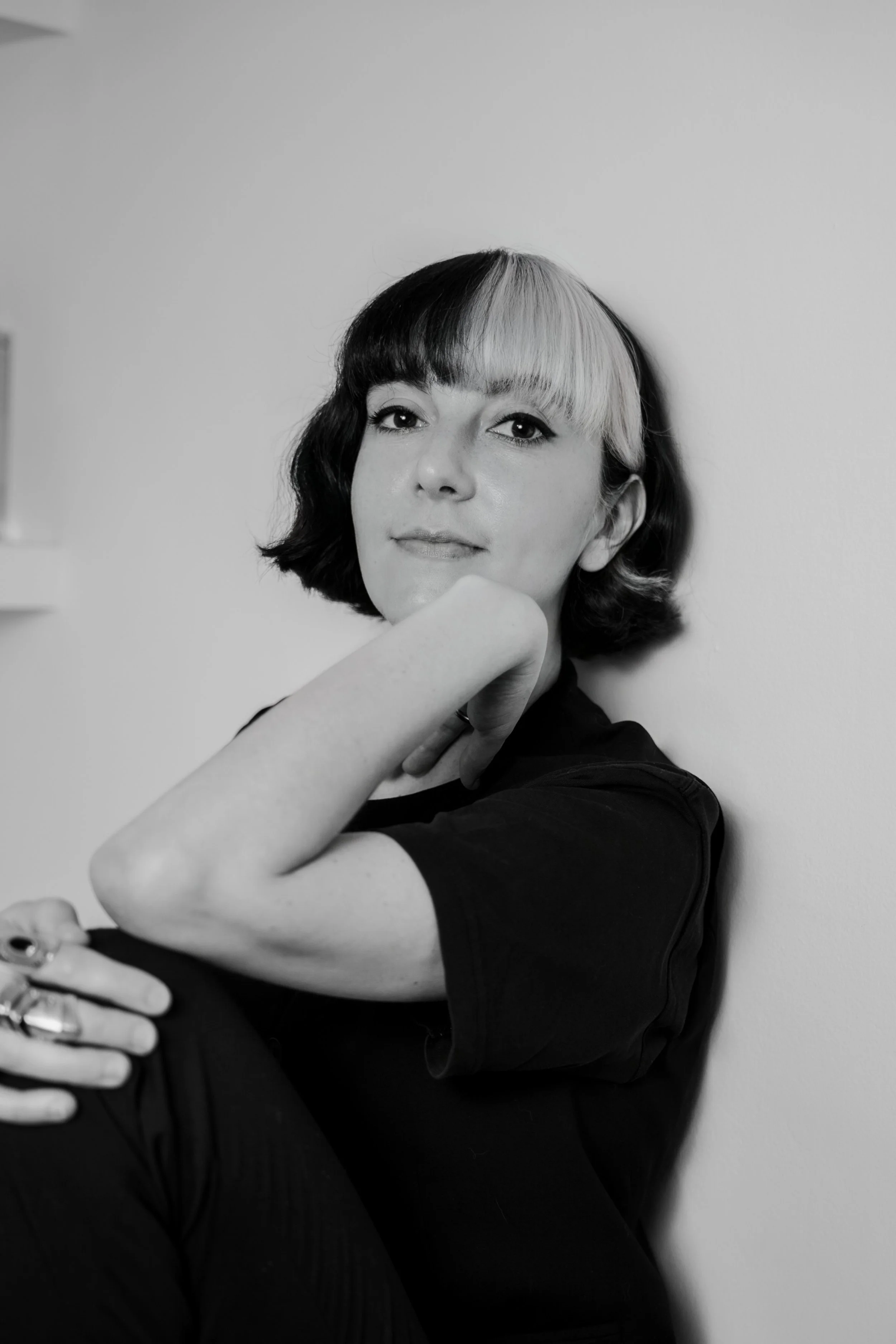 Black and white portrait of a woman with dual-colored hair, one side dark and the other light, posing with her hand resting on her chin against a plain wall.