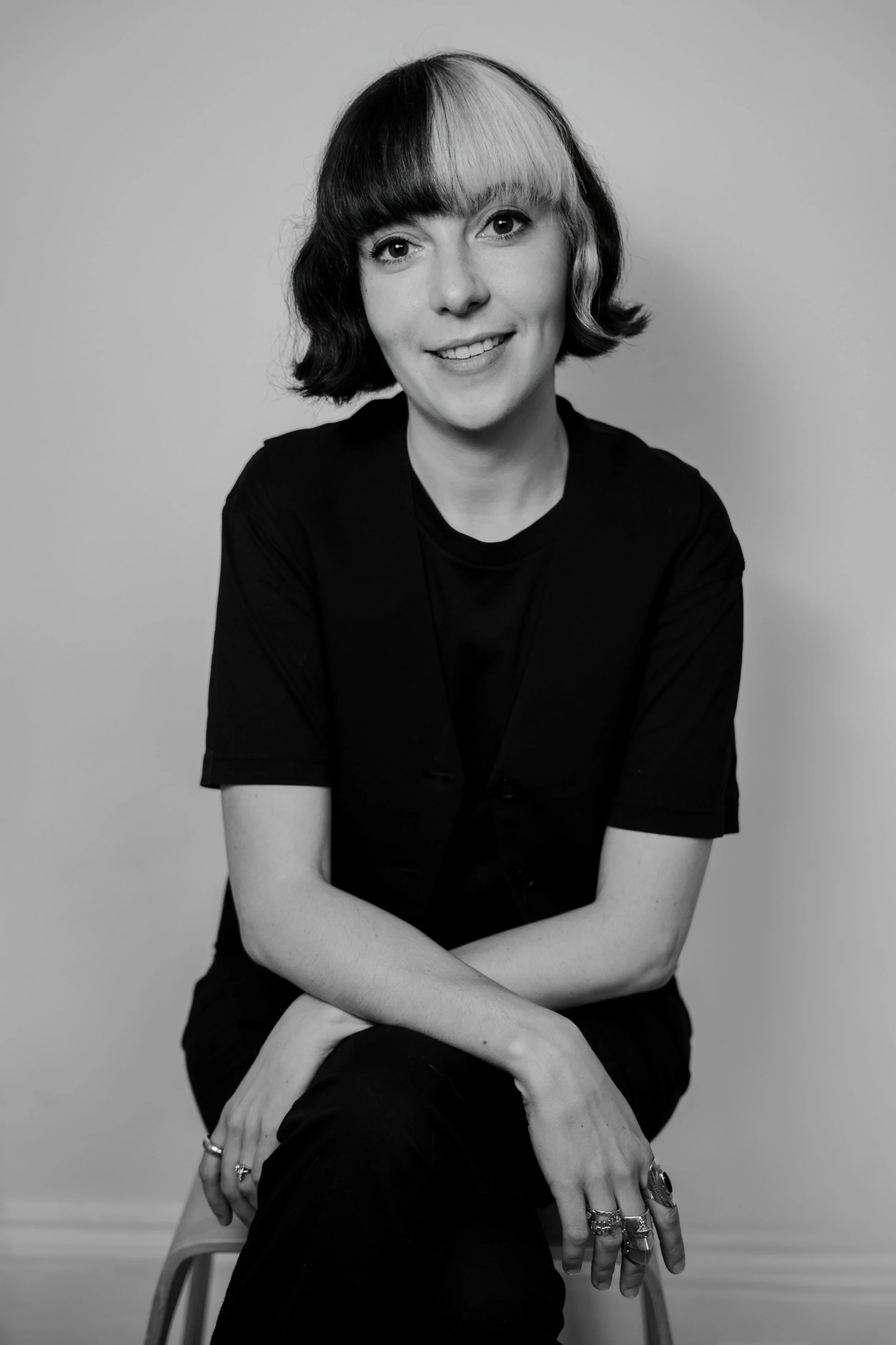 Black and white portrait of a woman sitting on a chair, wearing a dark shirt and multiple rings, with her arms crossed over her legs, smiling softly at the camera against a plain background.