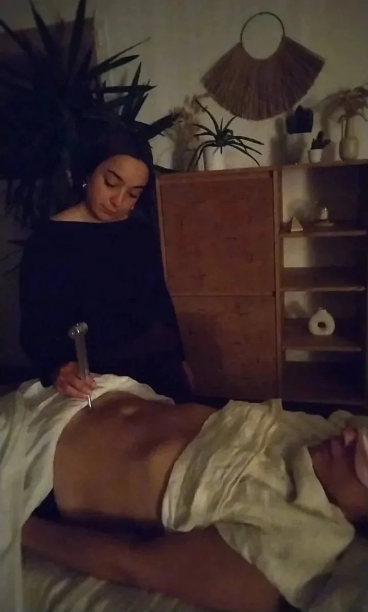 Praticienne réalisant un massage à l’aide d’un outil métallique sur le dos d’une personne allongée, dans une pièce chaleureuse décorée de plantes.