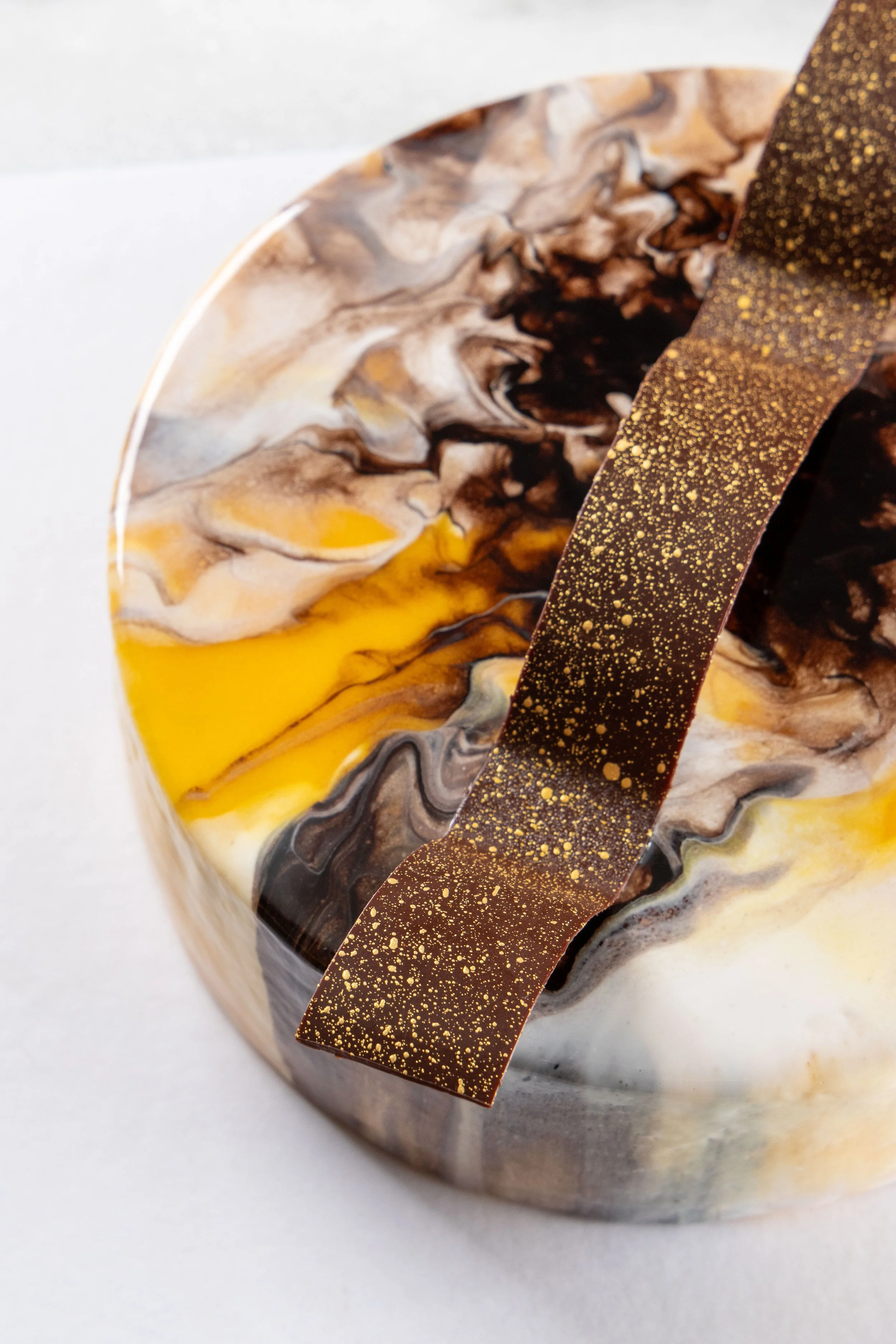Entremet Passion da Ahto Pâtisserie, com acabamento marmorizado em tons de chocolate, branco e amarelo e decoração de chocolate.