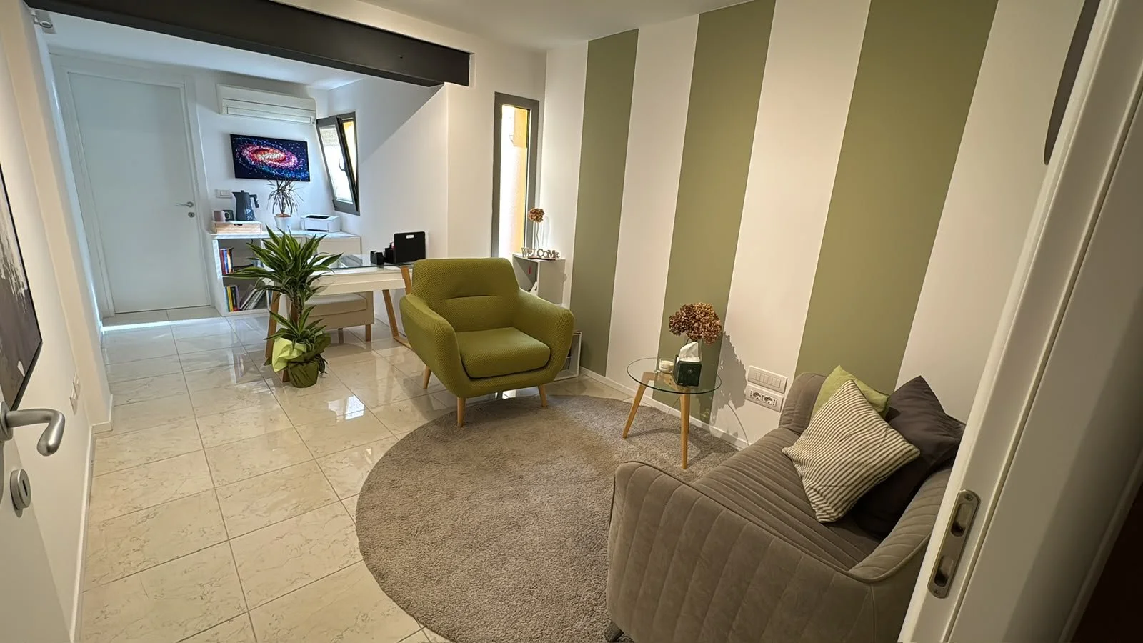 Interno di un soggiorno con divano beige, poltrona verde, tavolino con fiori, pareti con strisce verdi e bianche, piante e una zona con scrivania e schermo TV.