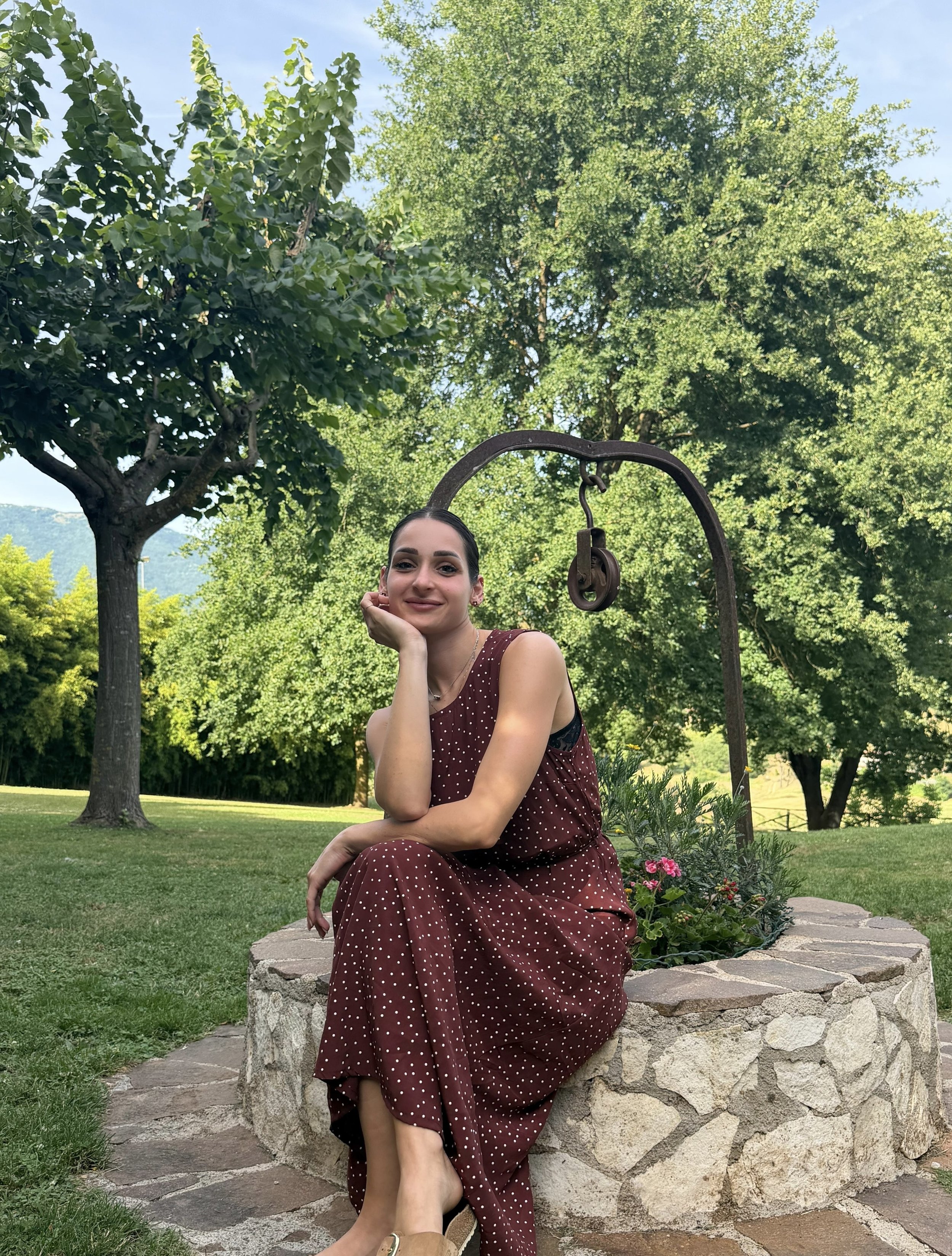 Una donna siede su una panchina di pietra in un giardino, indossa un vestito a pois marrone. Ricorda un giardino con molti alberi verdi e erba, sotto un cielo blu con qualche nuvola.