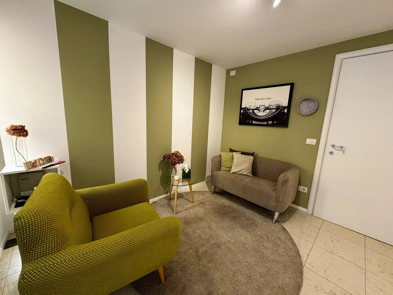 Soggiorno con divano verde, poltrona beige e pareti a righe verde e bianco, decorazioni, quadro in bianco e nero e orologio.