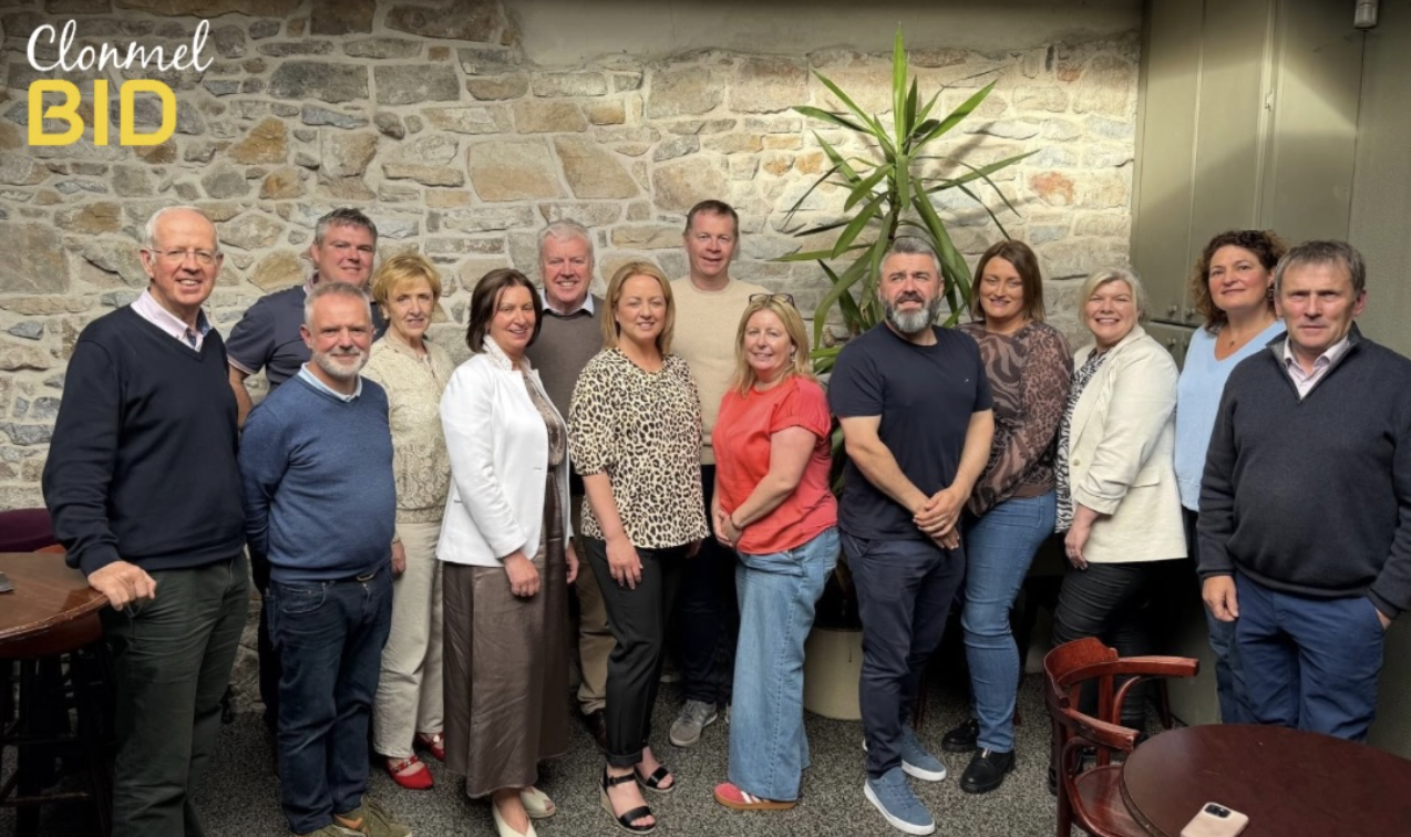 Group of fifteen people standing indoors in front of a stone wall, smiling at the camera, with a large green plant in the background.