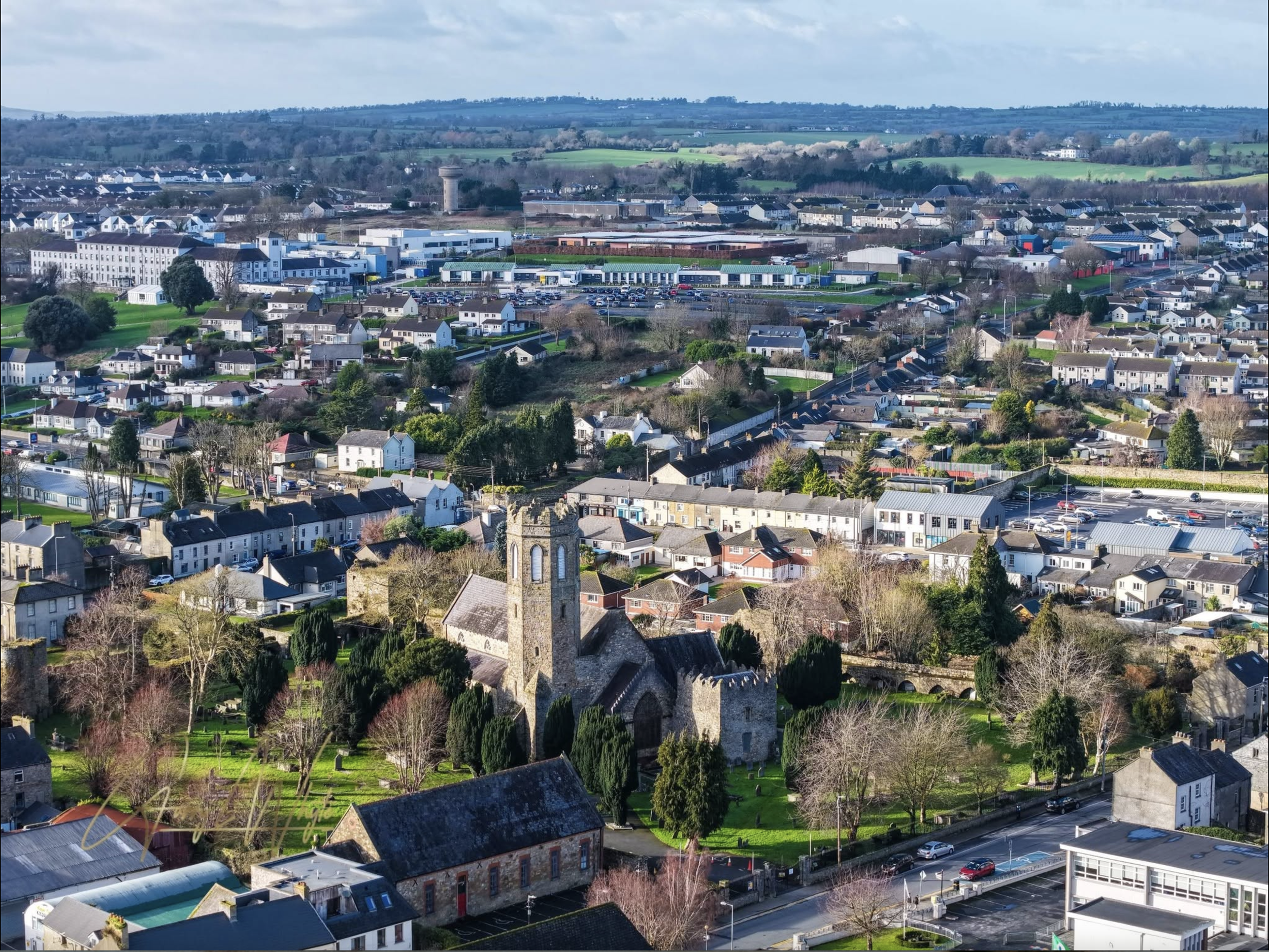 A small town with a mix of houses, trees, and commercial buildings, with a church in the foreground and rolling green countryside in the background.