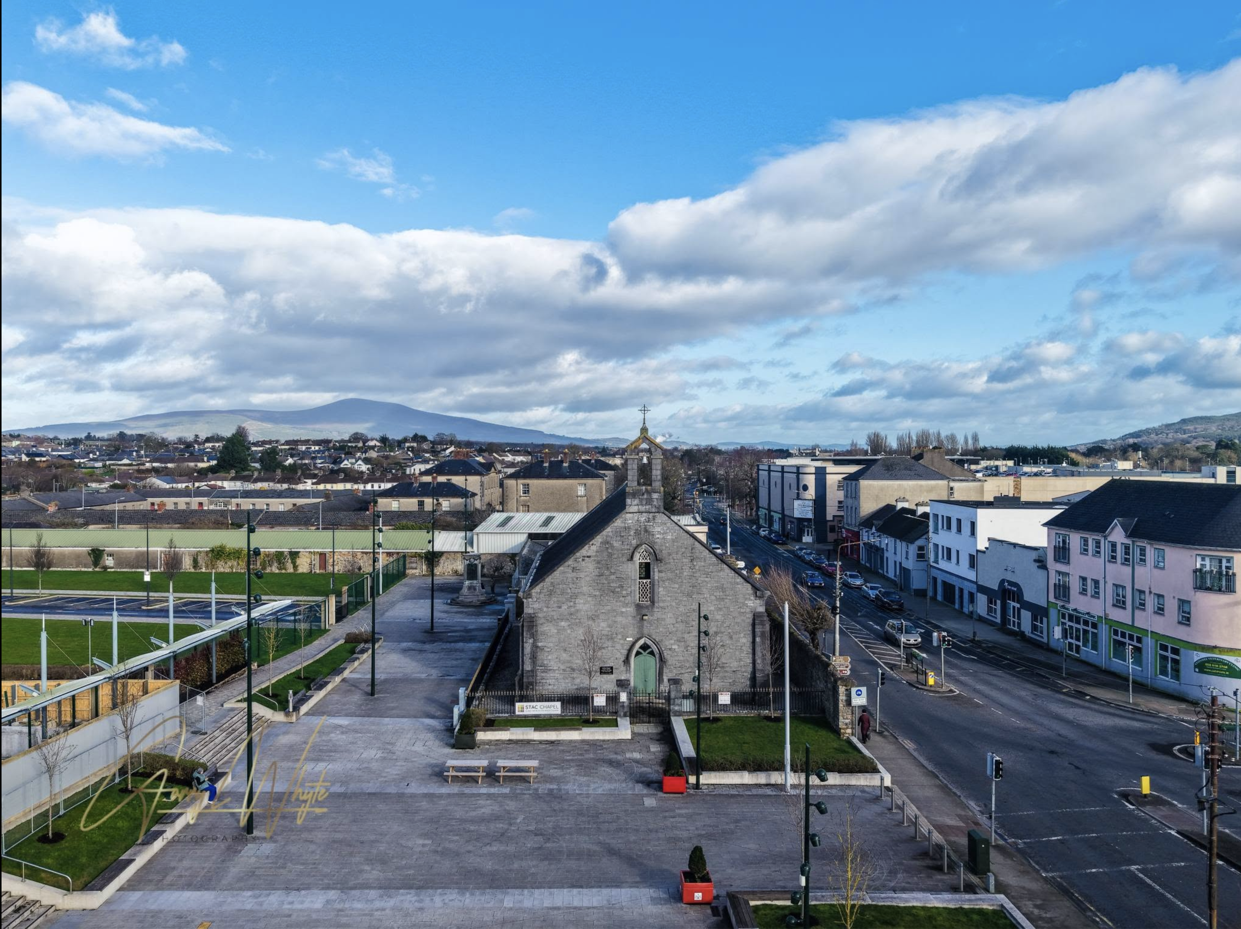 A cityscape view with a church, modern buildings, parking lot, and tennis court, under a partly cloudy sky with mountains in the background.