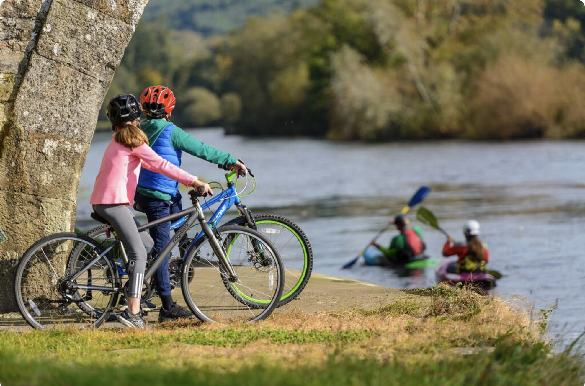 Children in helmets and jackets riding bicycles and kayaking by a riverside on a fall day.