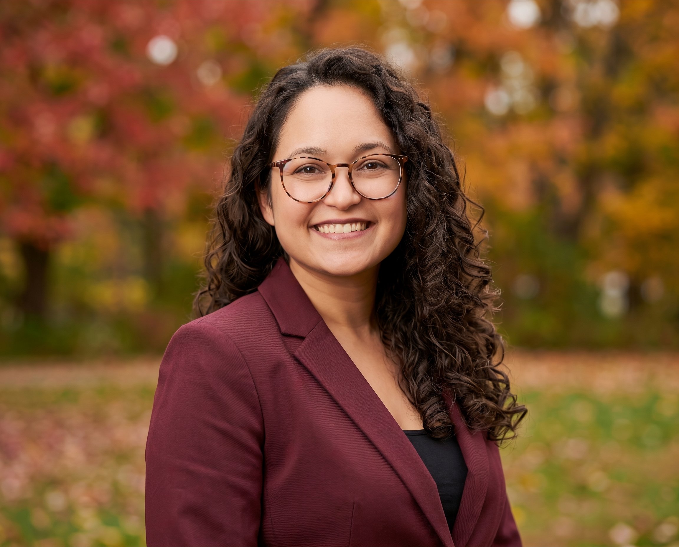 A woman with curly brown hair and glasses smiling outdoors with autumn colorful leaves in the background.