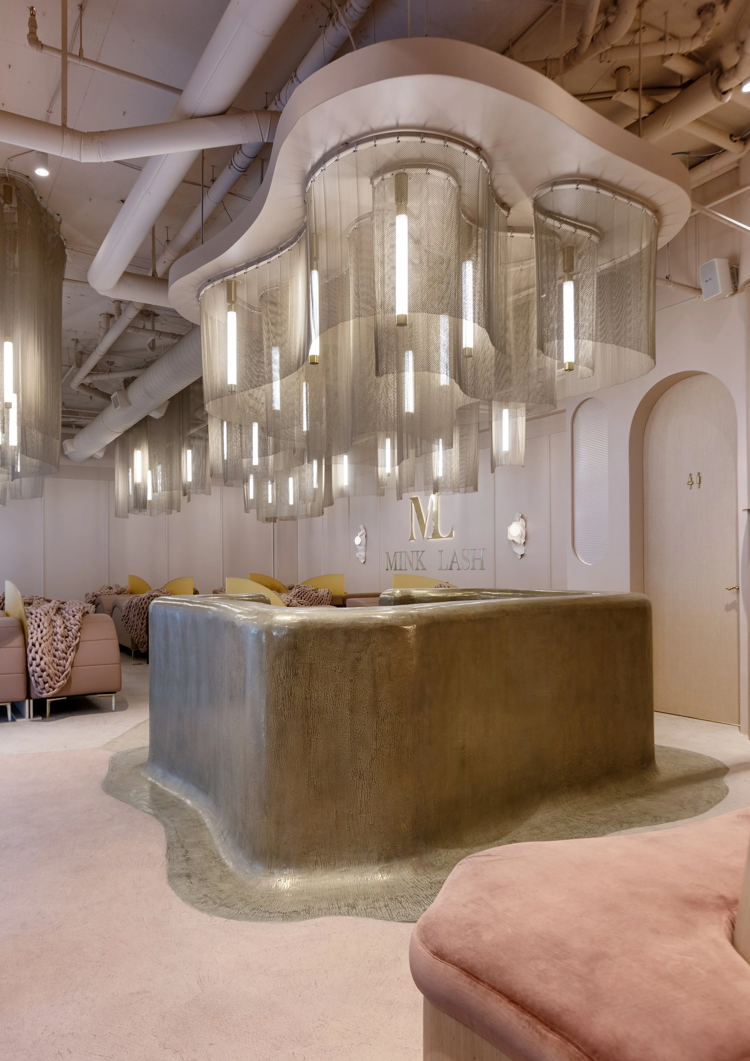Modern, stylish interior with soft pink and gold tones, featuring velvet couches, a gold metallic centerpiece table, and decorative gold lettering on the wall that says 'MINK LASH'.
