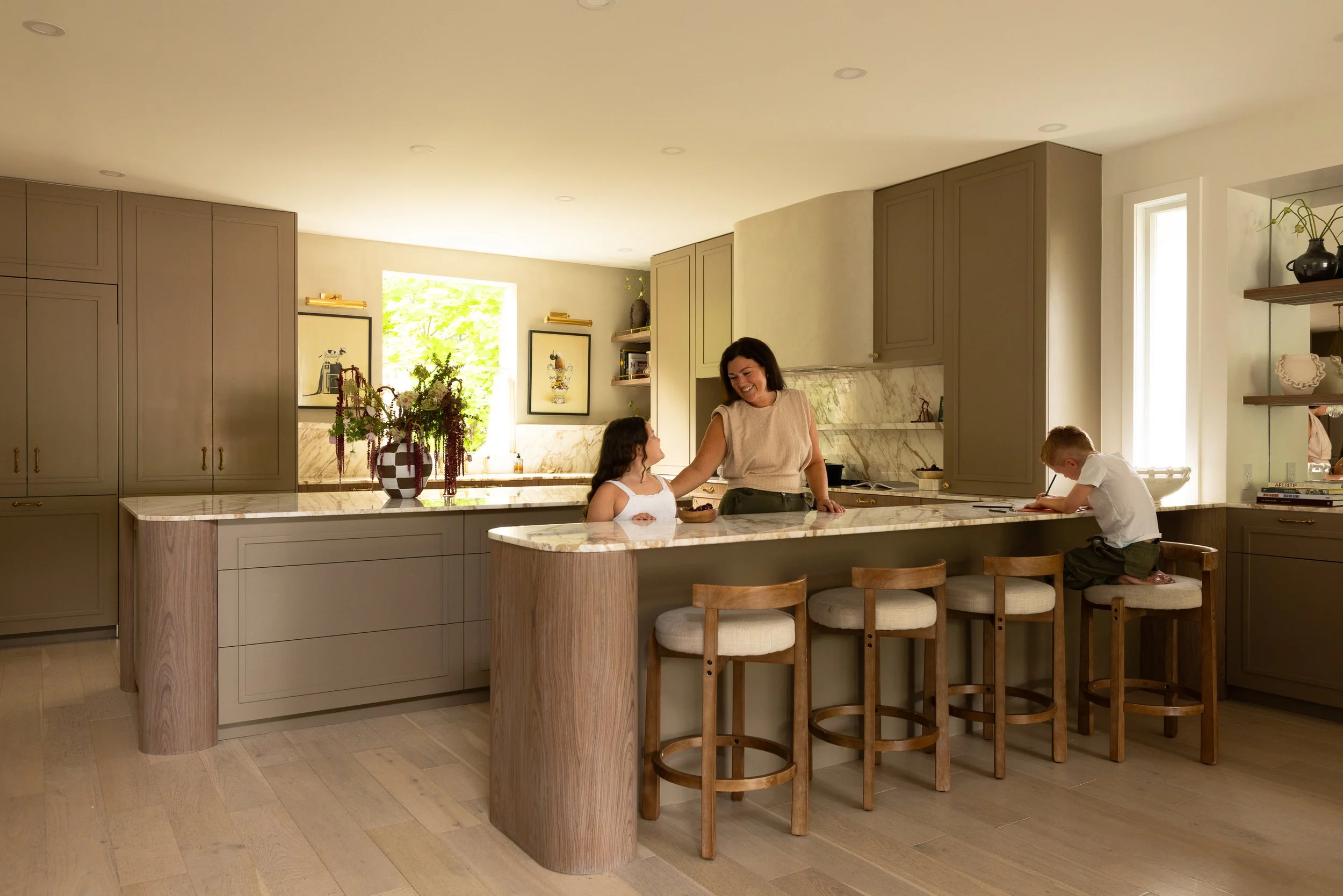 A bright kitchen with beige cabinetry and marble countertops, decorated with a large floral arrangement, featuring a woman and a young girl sitting at the counter, and a boy drawing on a stool.