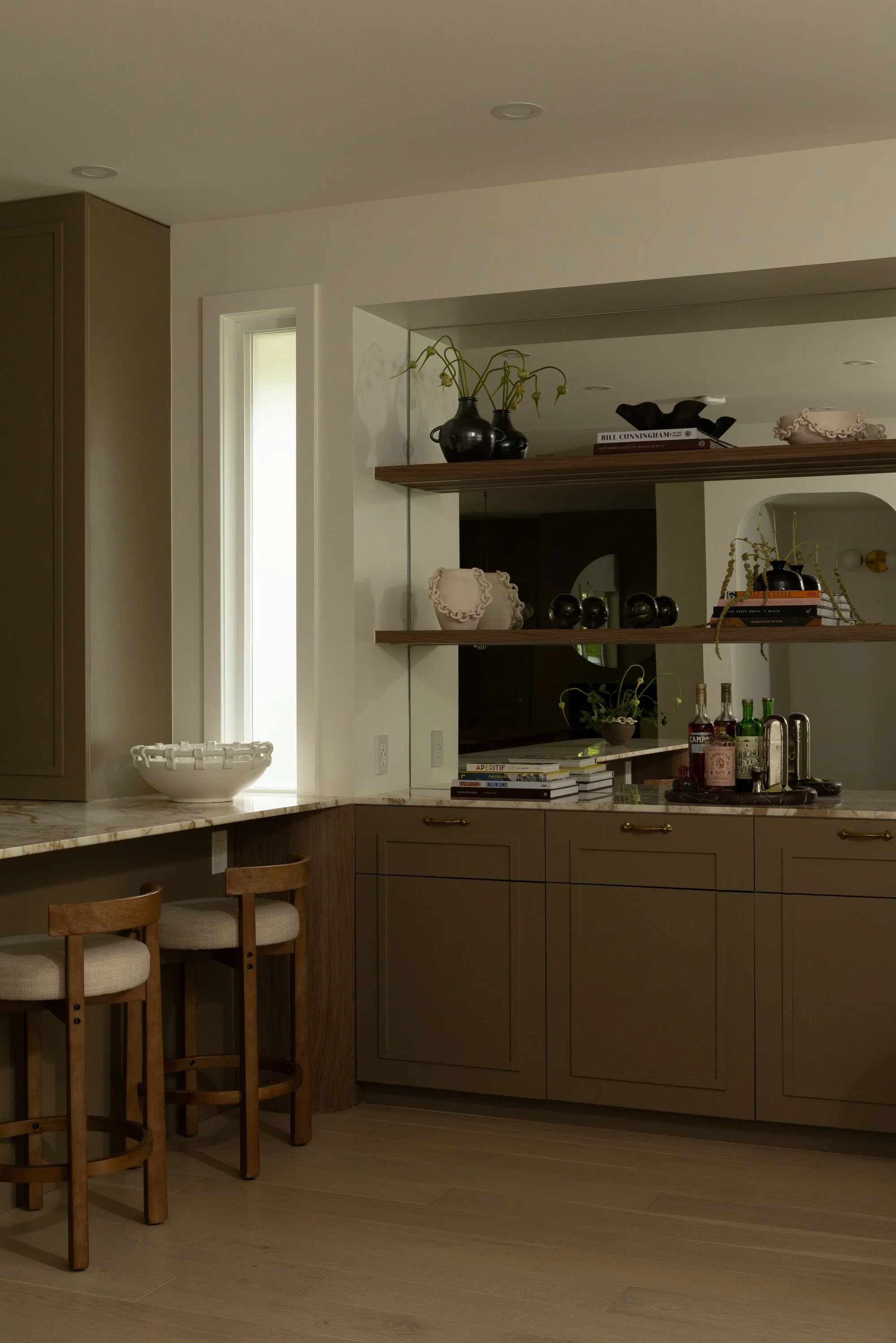 Modern kitchen with beige and wooden cabinetry, bar area with three stools, open shelves with decorative vases and books, and a marble countertop.