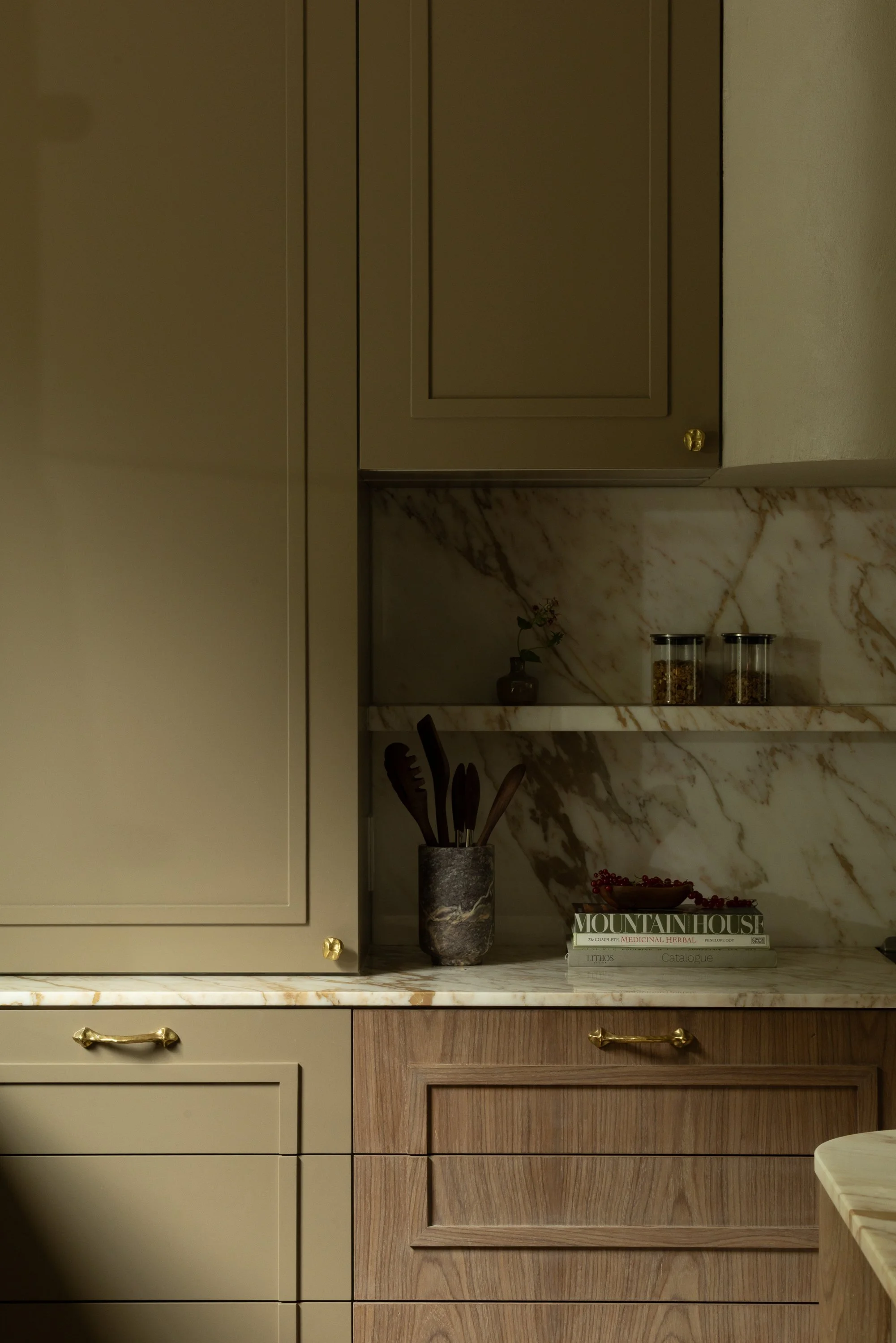 Close-up view of kitchen cabinets with beige and wood finishes, marble countertop, and decorative items including a potted plant, glass jars, and books.