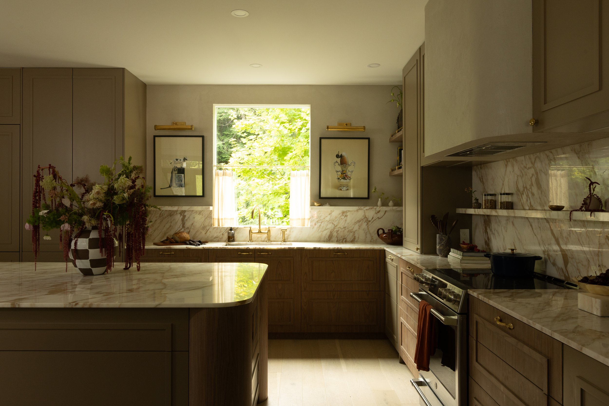 A modern kitchen with wooden cabinets, marble countertops, and a large window letting in natural light. Decor includes framed art, a bouquet of flowers, and various kitchen utensils.