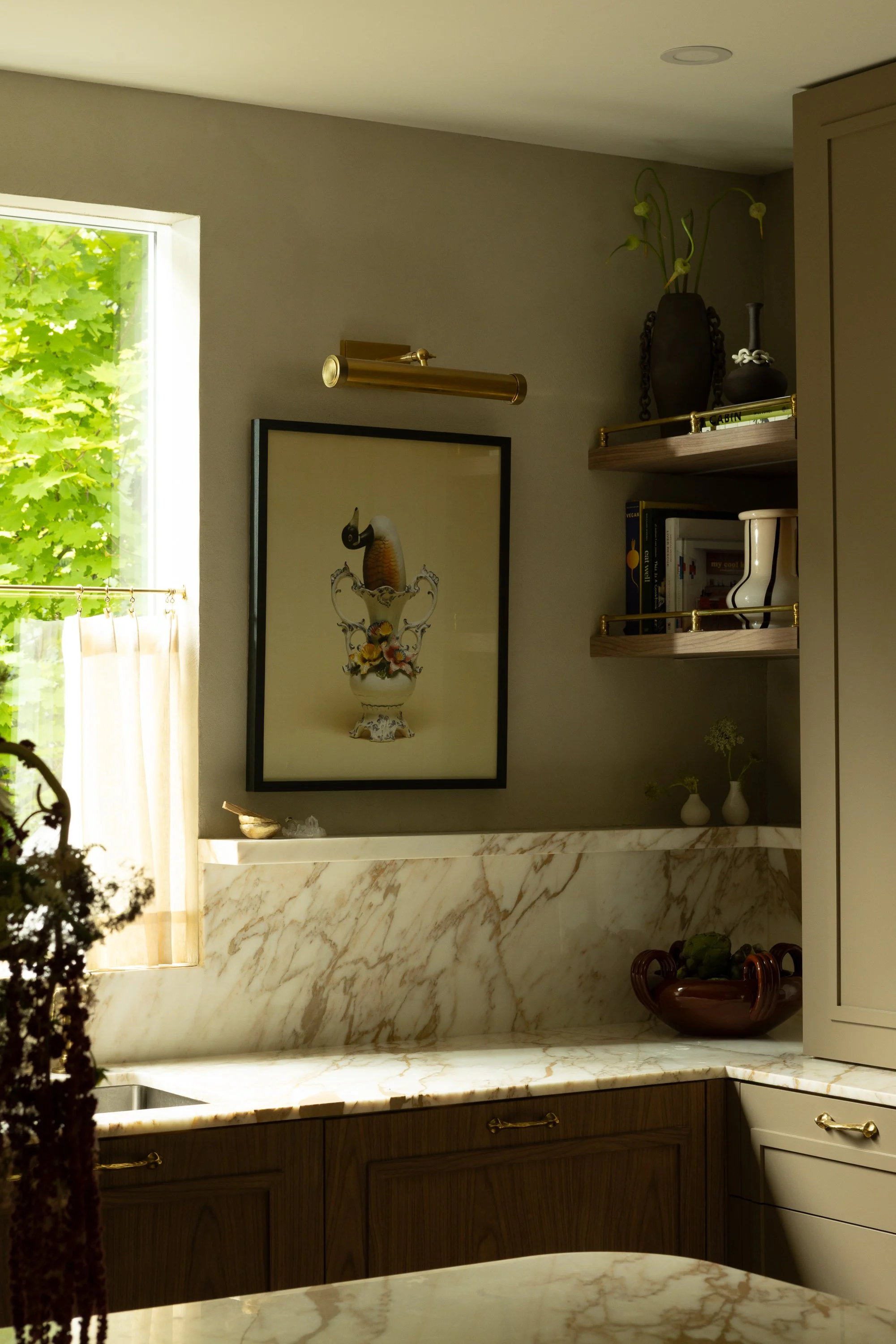 Modern kitchen with marble countertops, a wall-mounted brass light, framed artwork, a black vase with plant, and bookshelf with books and decorative items.