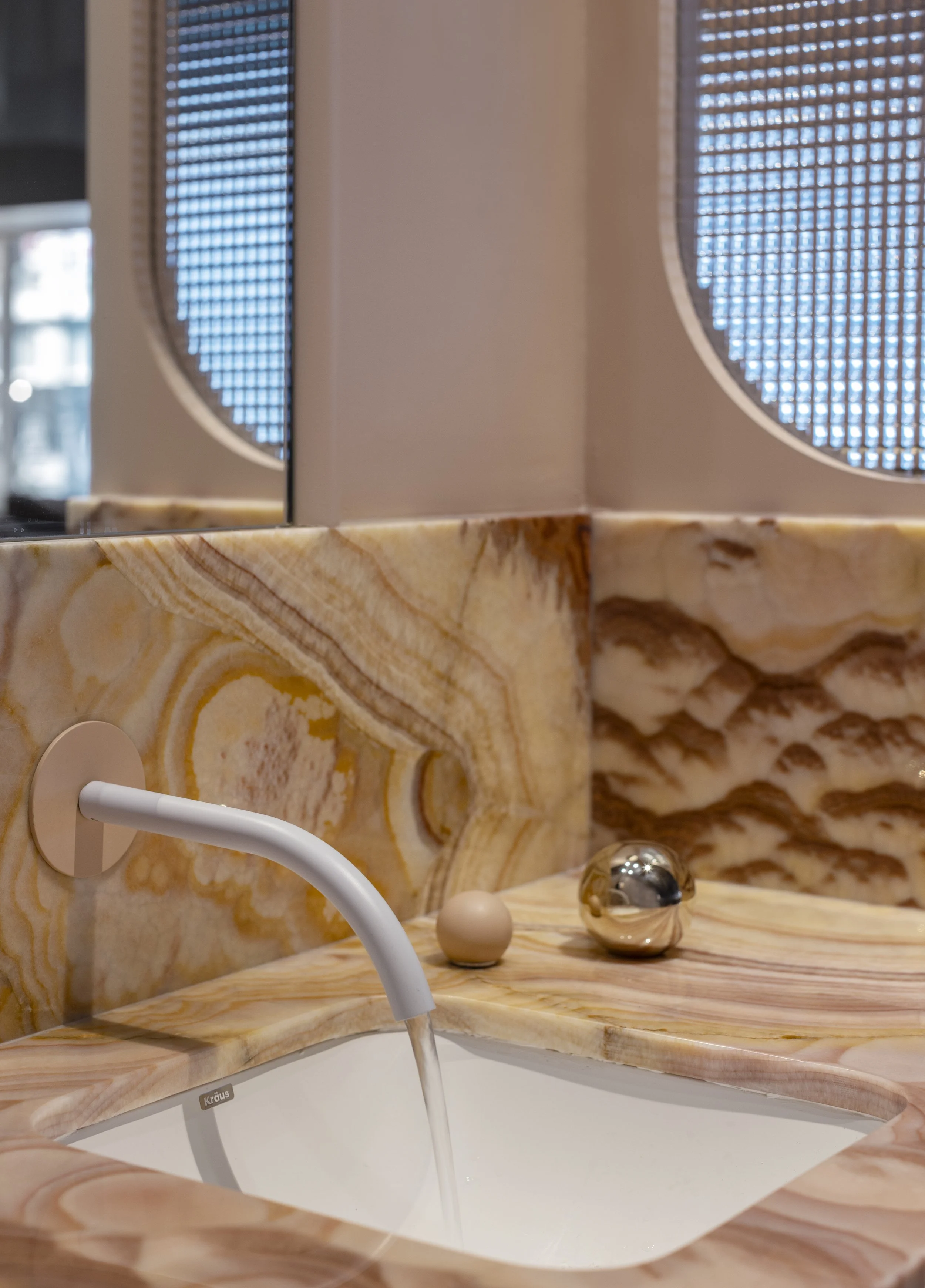 Close-up of a marble bathroom sink with gold, beige, and brown swirls, white faucet, and a mirror reflecting a window with blue patterned blinds.