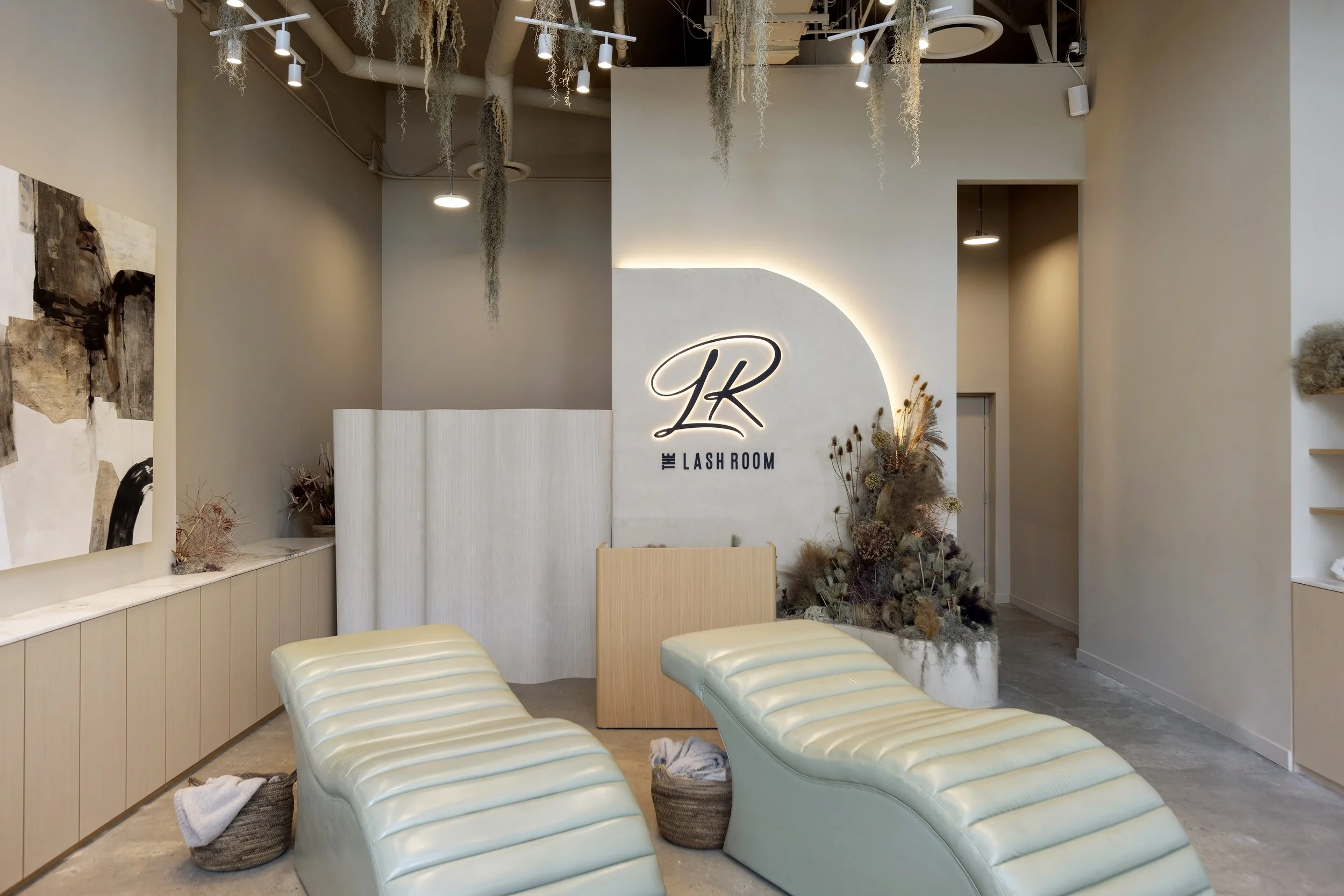 Waiting area with two cream-colored lounge chairs, wicker baskets with towels underneath, and dried plants near a wall with the sign 'The Lash Room.' Modern decor with hanging plants and artwork on the wall.