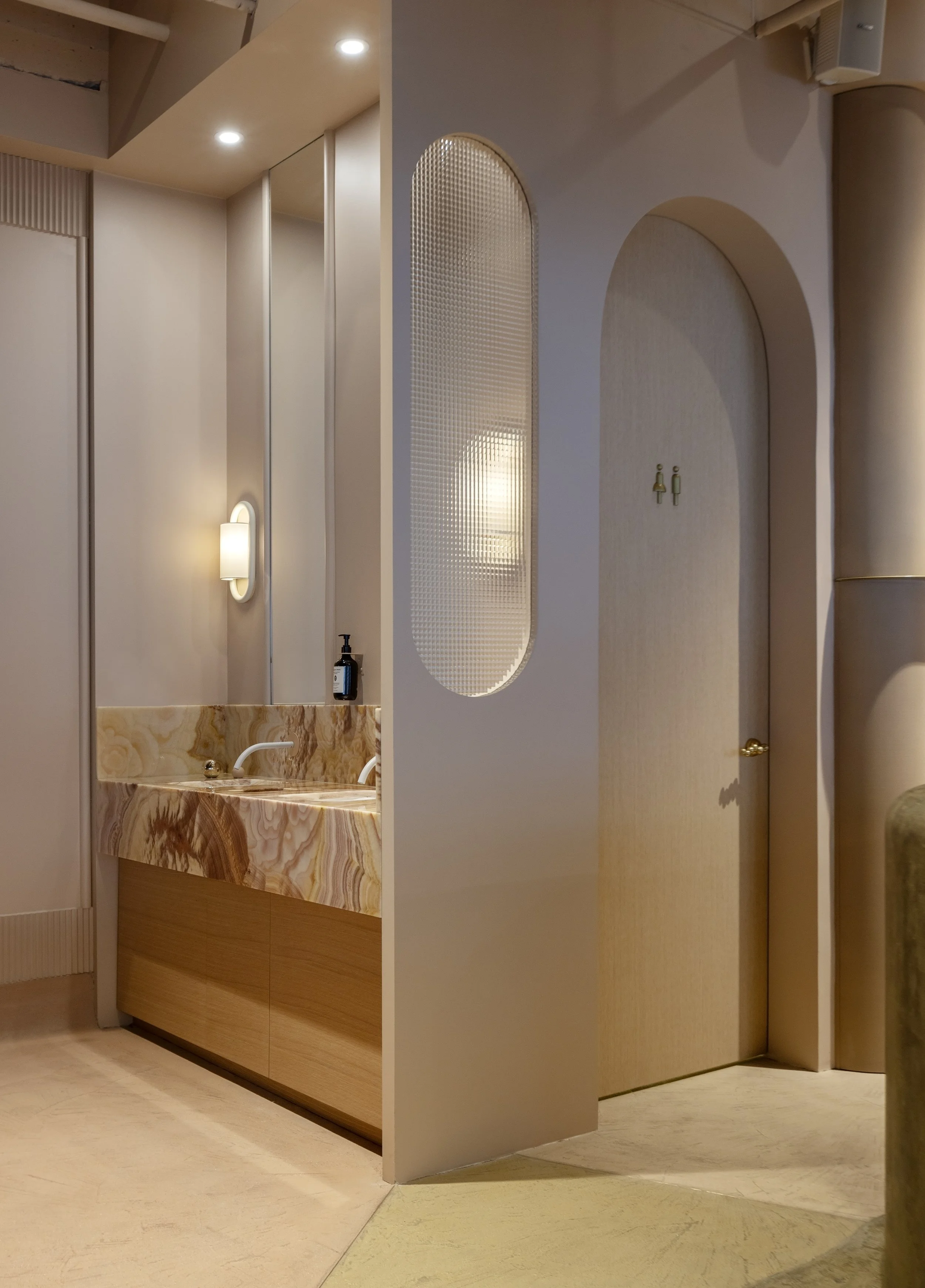 Interior of a modern bathroom with a marble vanity, wall-mounted light, and a partition wall with a textured oval window, and a toilet area in the background.
