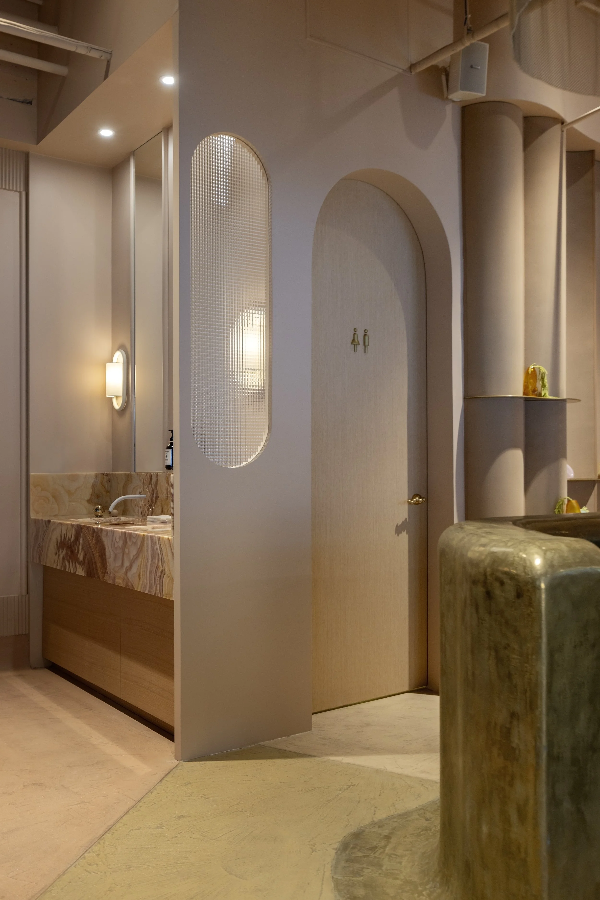 Interior of a modern space with beige walls, a circular marble counter in the foreground, a small sink with a marble countertop, and a decorative door with a gold handle. The room features subtle lighting and architectural details like curved walls and columns.