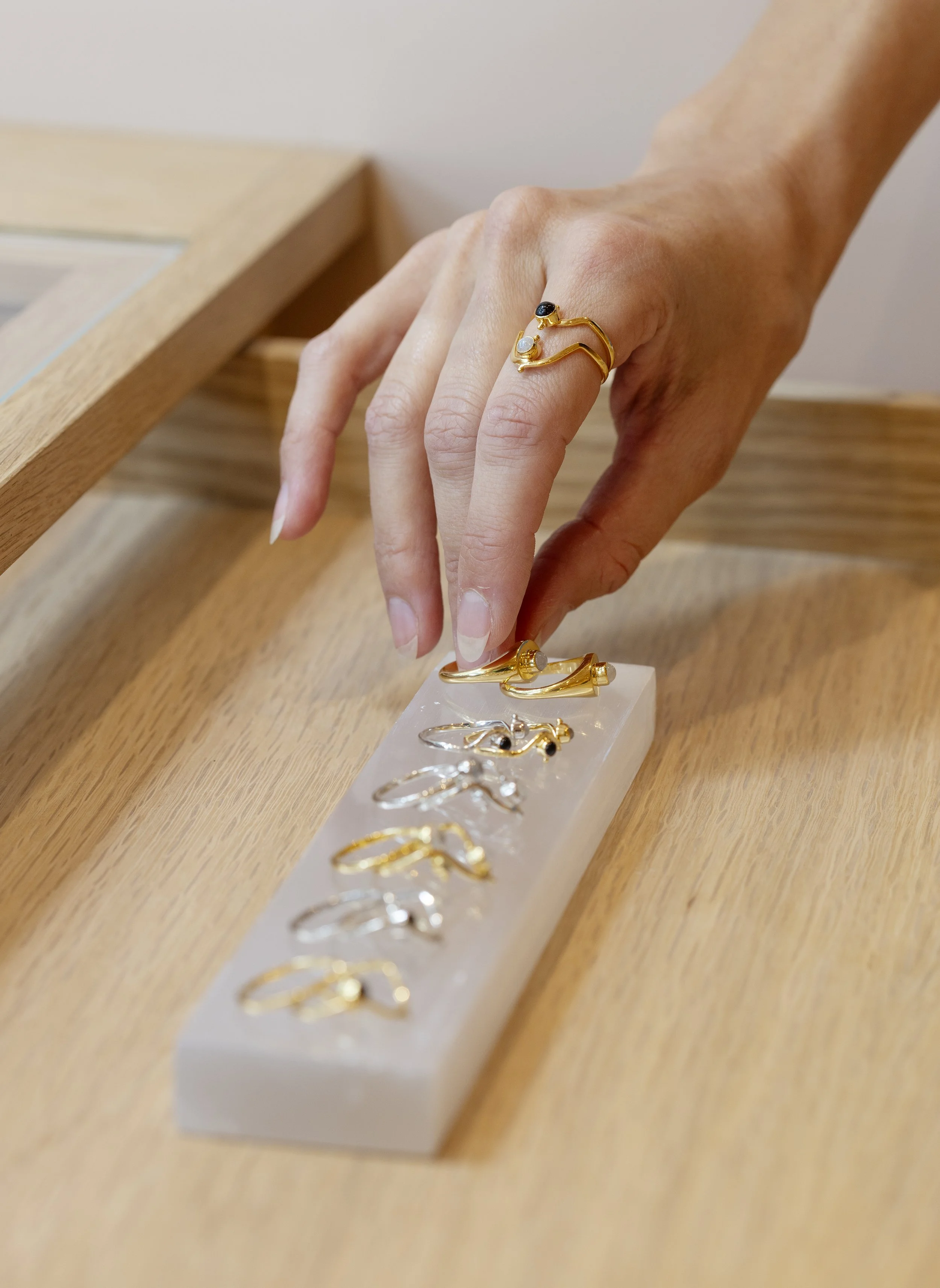 Close-up of a hand wearing a gold ring with black and white stones, resting on a wooden display tray with other gold rings. The tray is on a wooden surface.