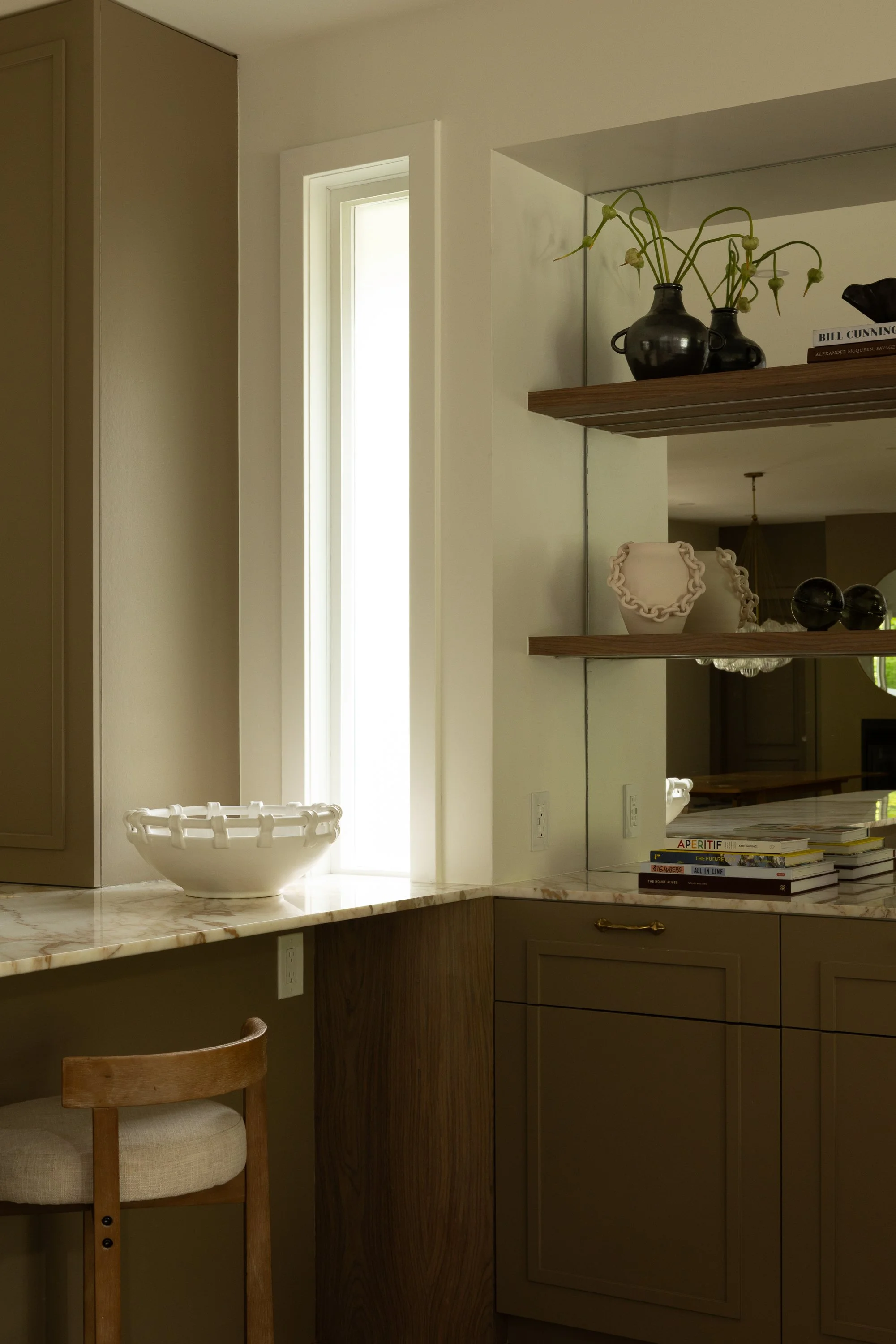 A kitchen with gray cabinets, a marble countertop, a large window, a decorative white bowl, a stack of books on the counter, and hanging shelves with black vases, books, and decorative objects.