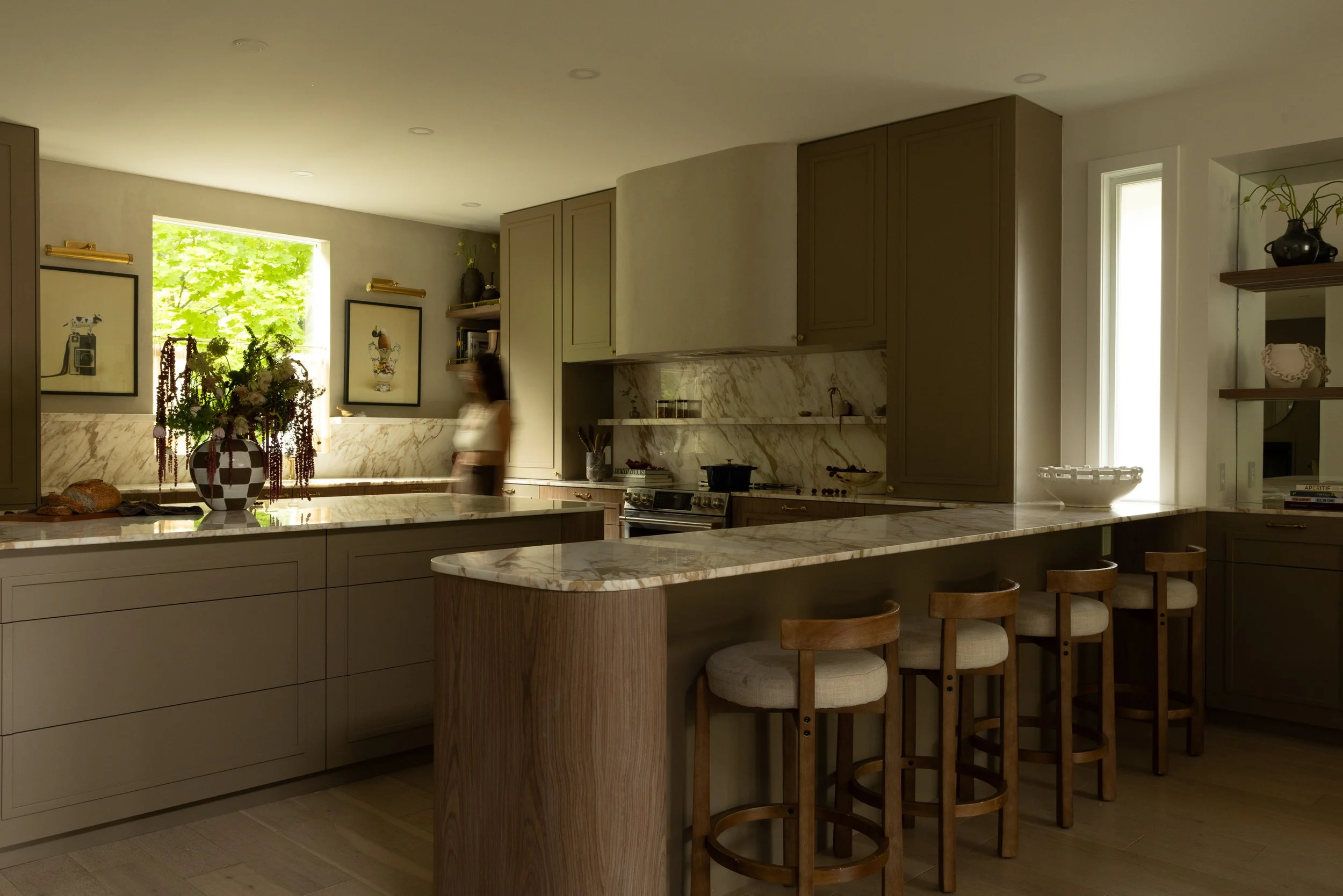 A modern kitchen with beige cabinets, marble countertops, and a breakfast bar with wooden stools. There is a person blurry in motion near the window, which has green foliage outside. Decor includes framed artwork, vases with plants, and small decorative objects.