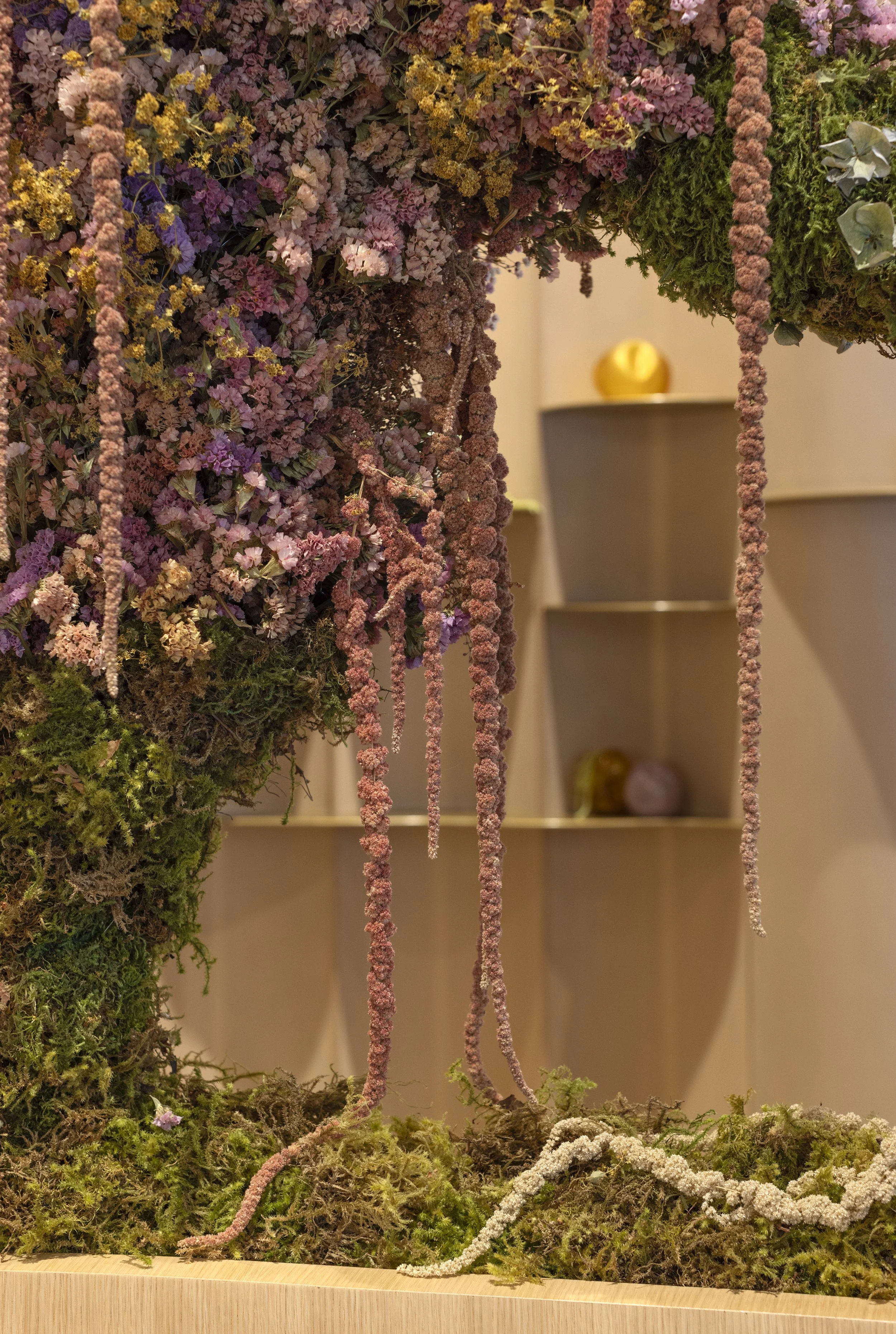 Arrangement of pink, purple, yellow, and white dried flowers and green moss hanging from a display with a light-colored wooden base, with shelves and golden decor visible in the background.