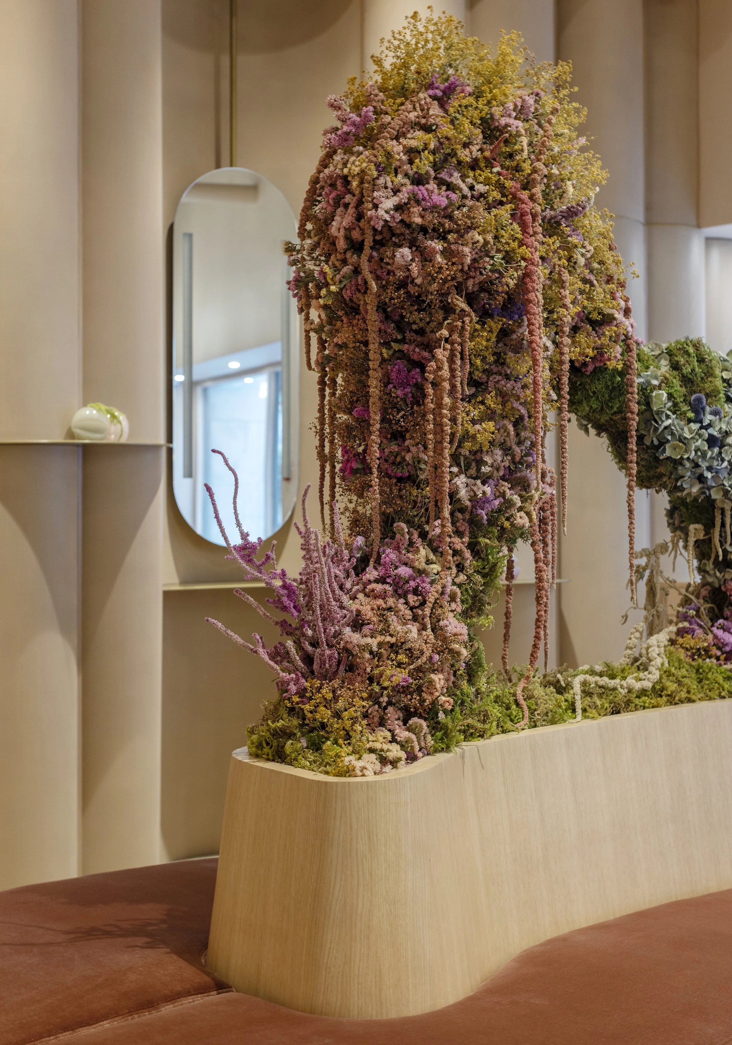A floral arrangement with pink, yellow, and purple flowers on top of a wooden table in a room with beige walls and a mirror.