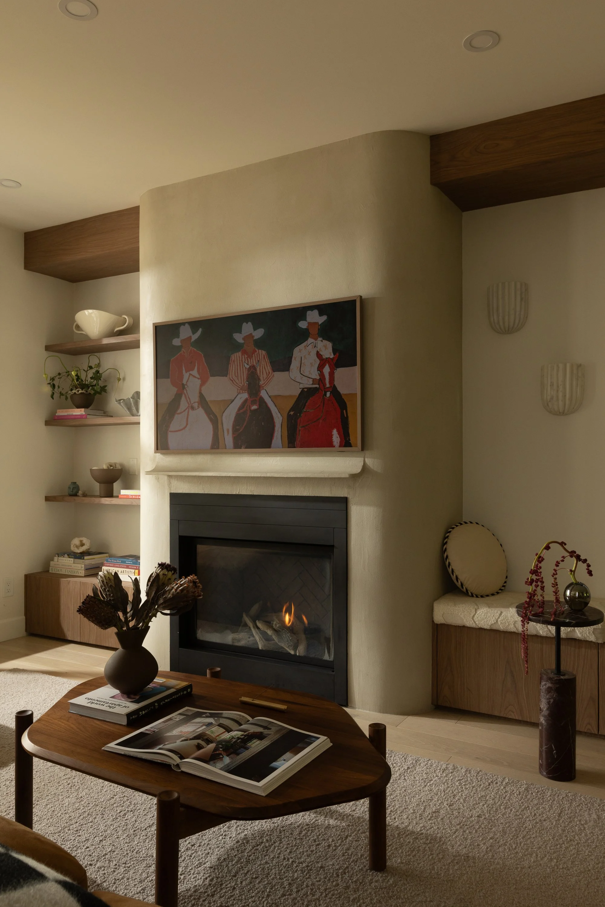 Living room with a wood coffee table, a black fireplace, a painting of cowgirls on the wall above the fireplace, and built-in shelves with decorative items.