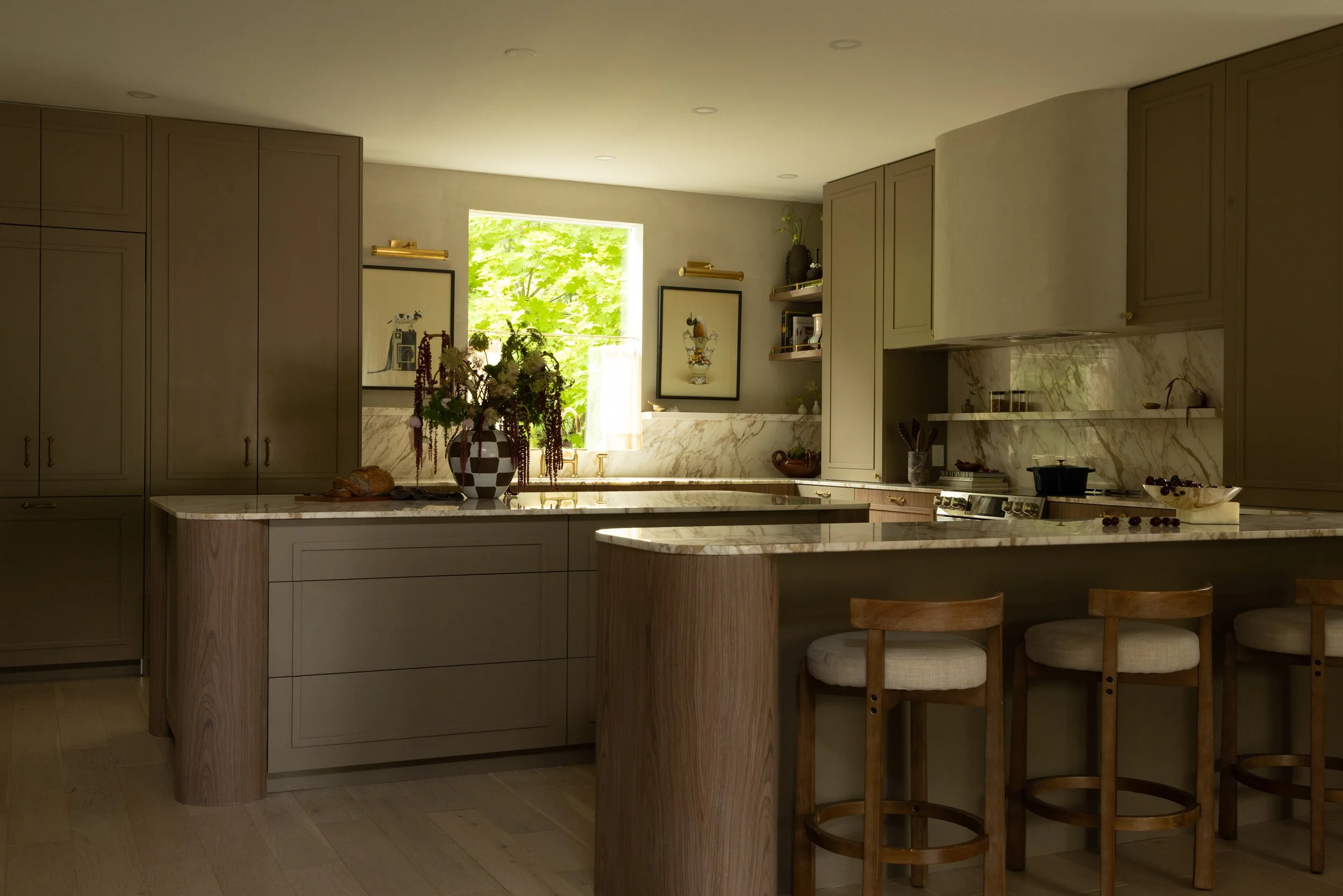 A modern kitchen with cream-colored cabinets, marble countertops, a window with green trees outside, decorative framed artwork, and a vase with flowers on the counter.