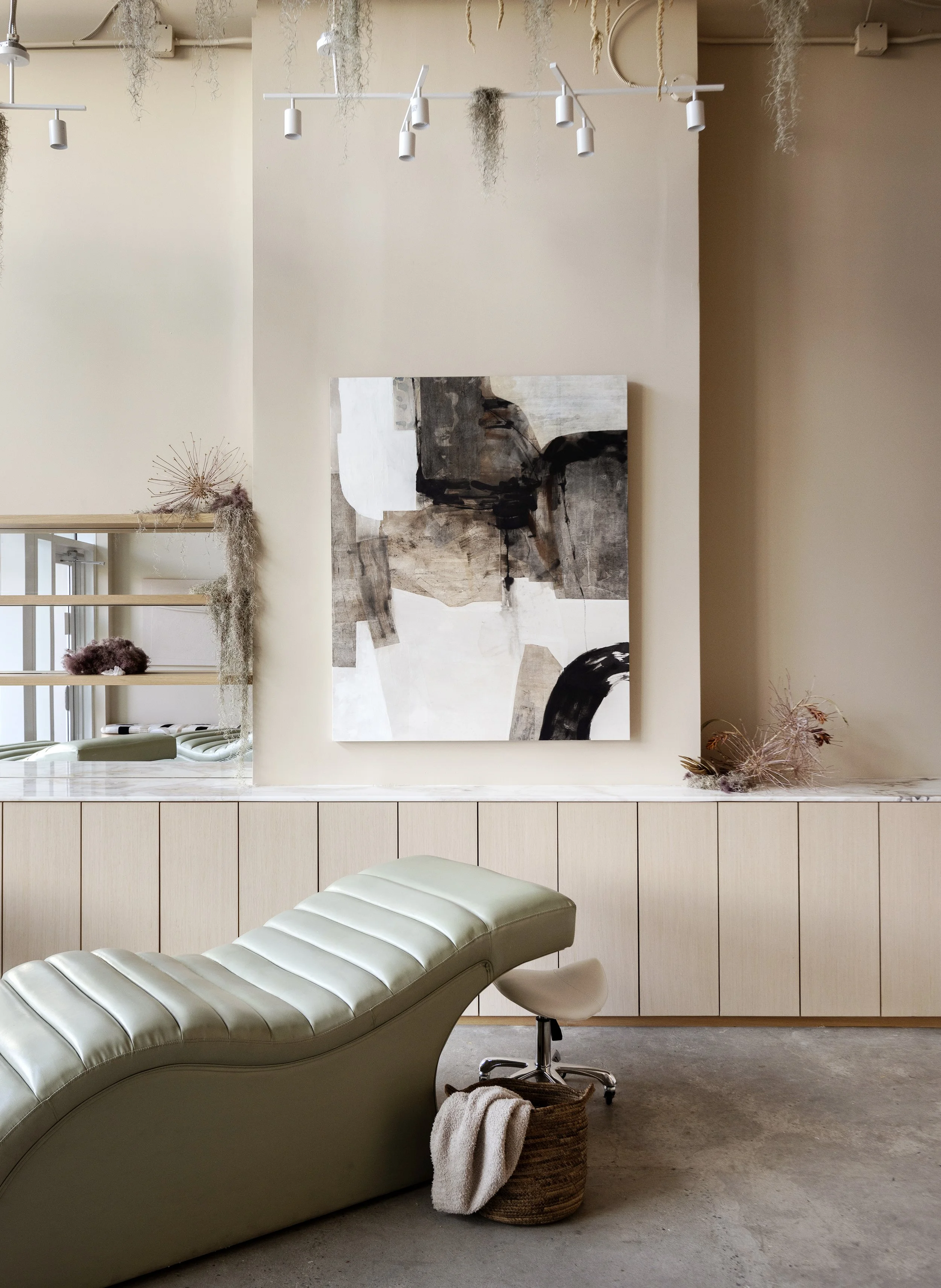 A modern, minimalist interior with a curved, beige lounge chair in the foreground, a small draped towel in a wicker basket beside it, modern furniture and artwork on the wall, and soft neutral color palette.