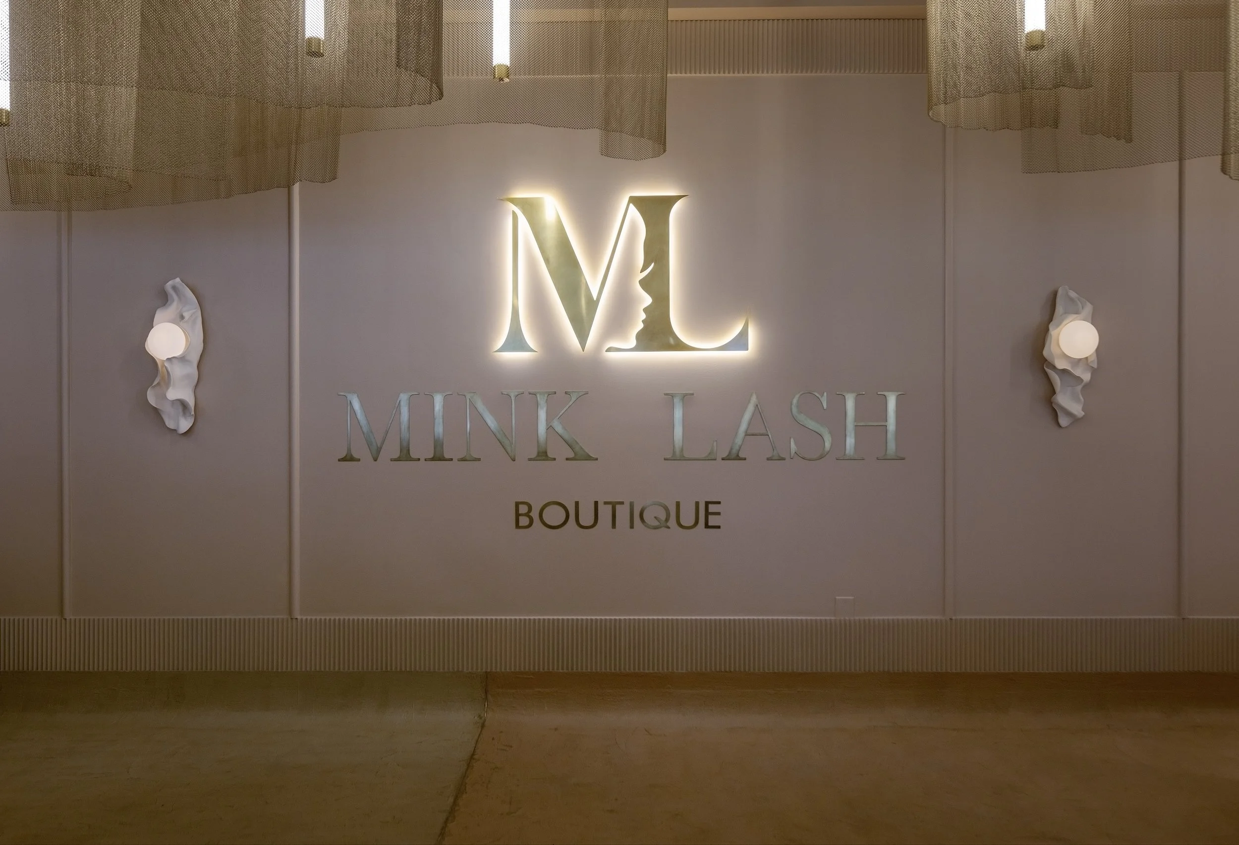 Illuminated sign for Mink Lash Boutique with a stylized 'M' and 'L' and a profile of a woman's face in the logo, flanked by decorative wall sconces with fabric shades.