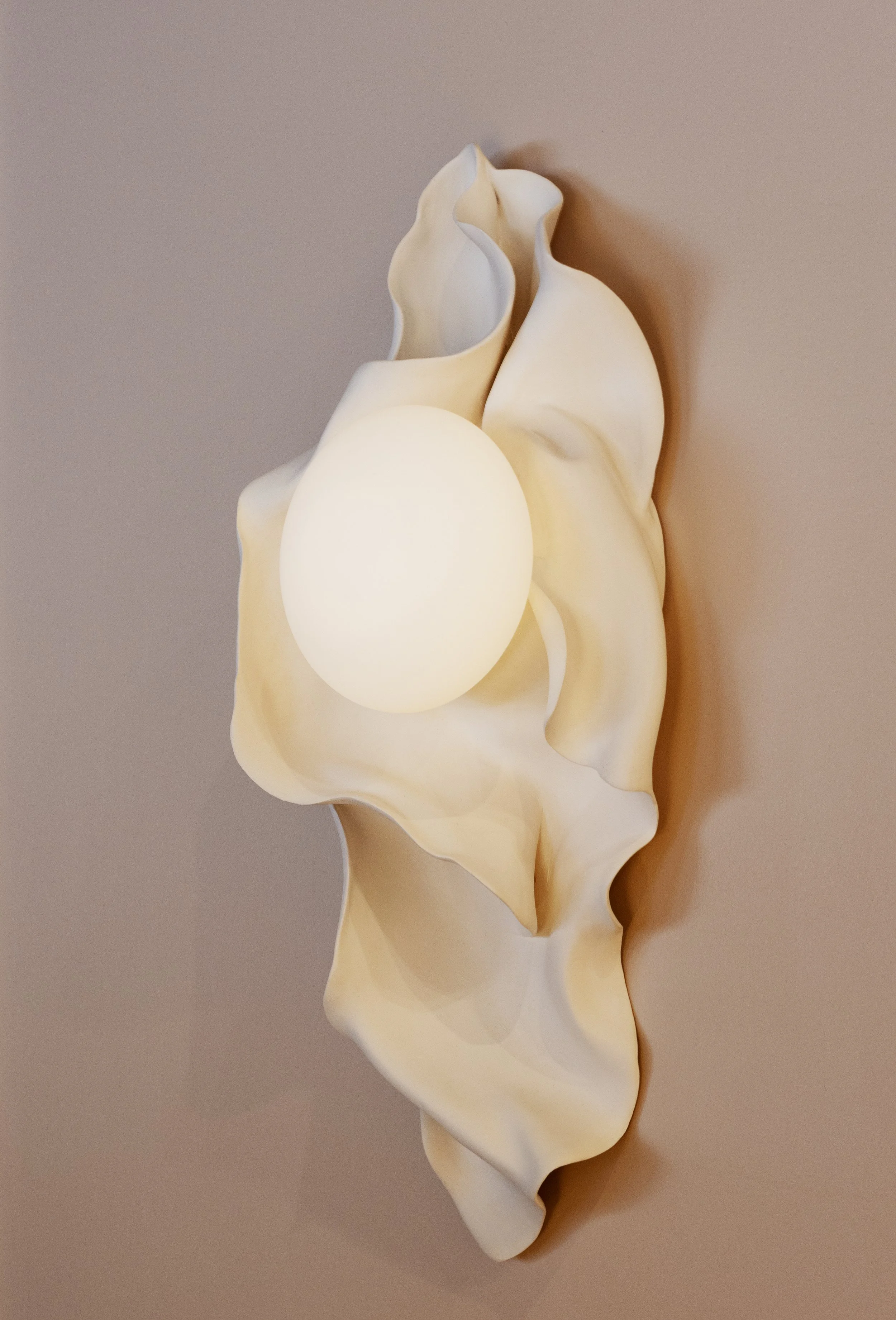 A white ceramic sculpture resembling a seashell with an abstract, organic shape, mounted on a neutral background.