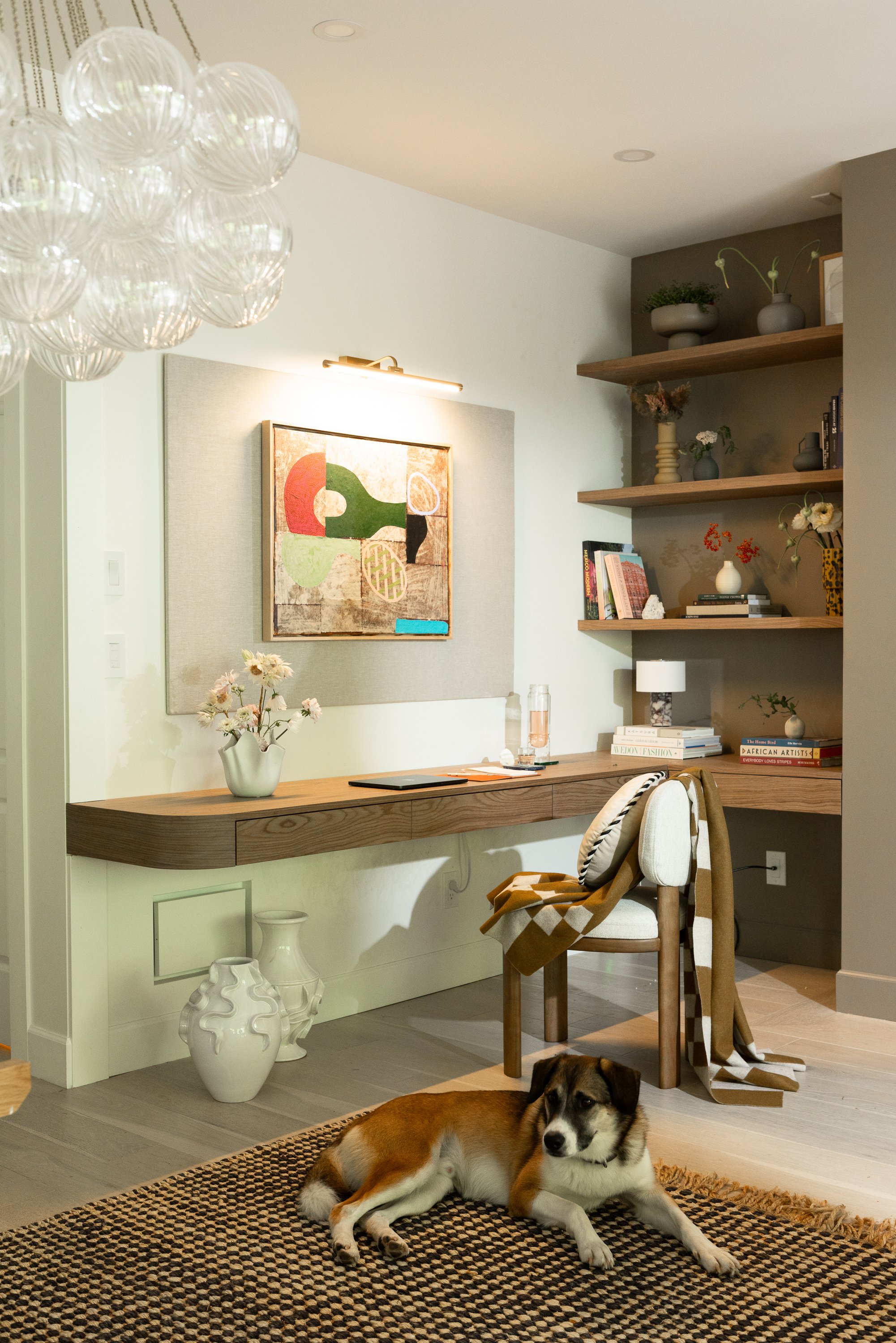 A cozy living room with a wooden desk, books, and decorative vases on floating shelves. A dog is lying on a patterned rug in front of the desk, with a beige and white chair nearby. White vases on the floor and a colorful abstract painting on the wall add to the decor.