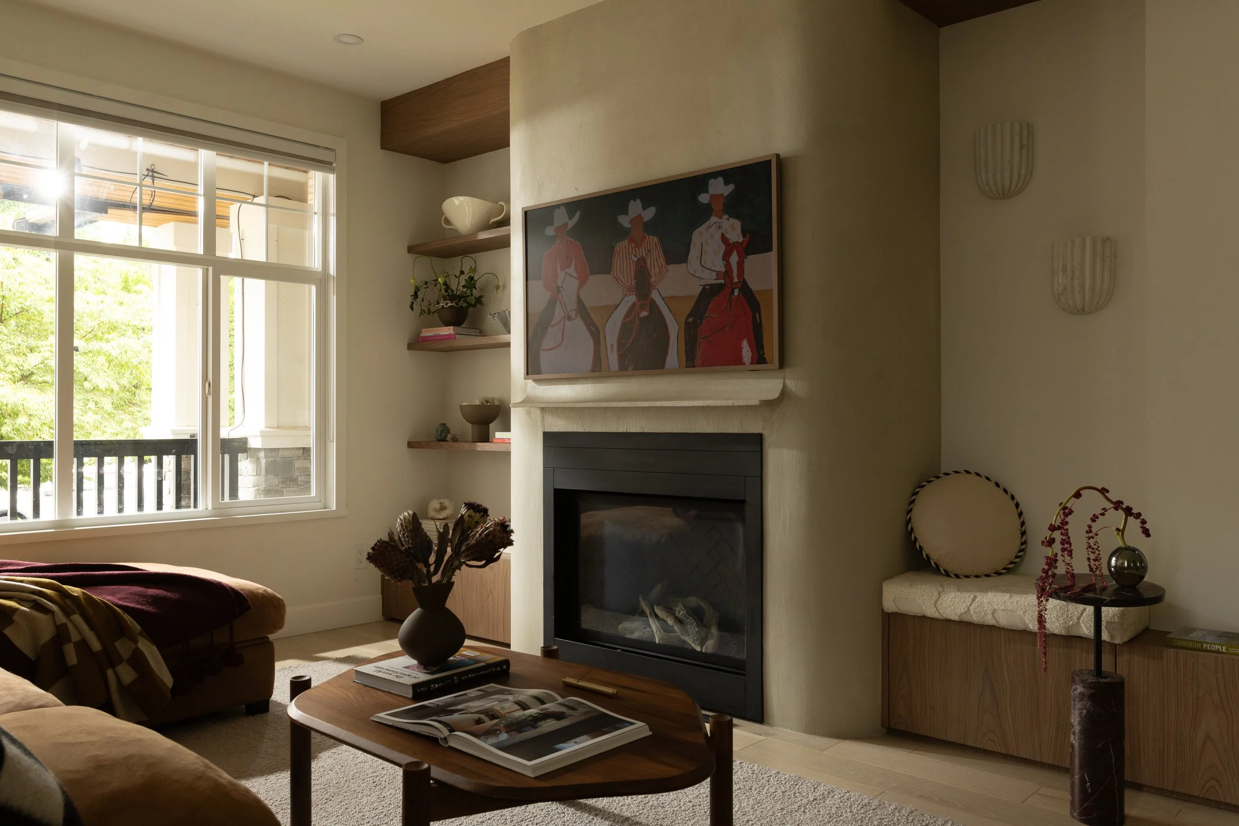 Living room with a window, a fireplace, wall art depicting cowboys, a chair, a side table with a vase and plant, and a coffee table with books.